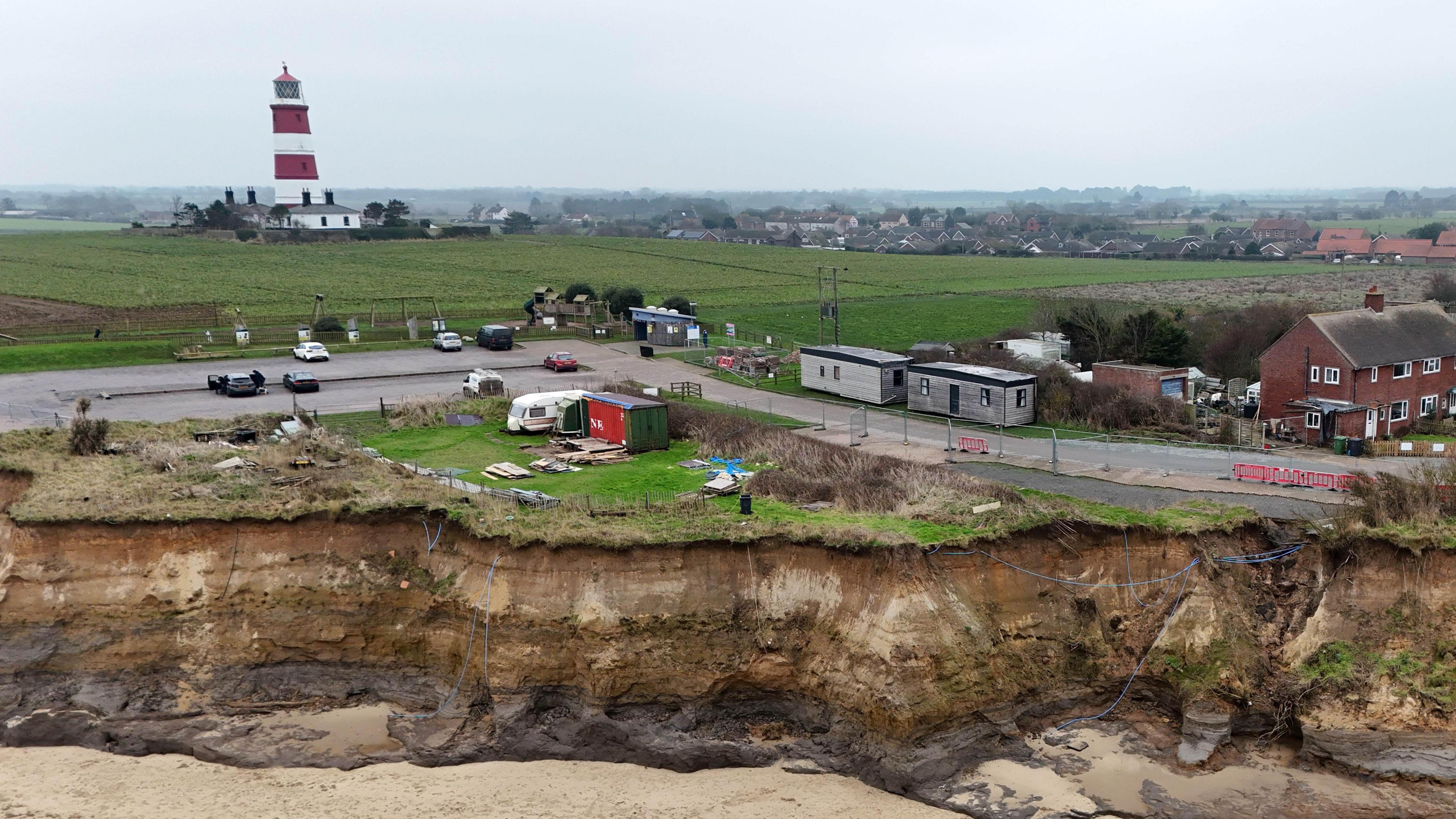 An aerial shot looking from above the sea towards the cliffs at Happisburgh. There is fencing on the road leading to the car park, preventing access to the crumbling cliffs. The lighthouse is in the background.