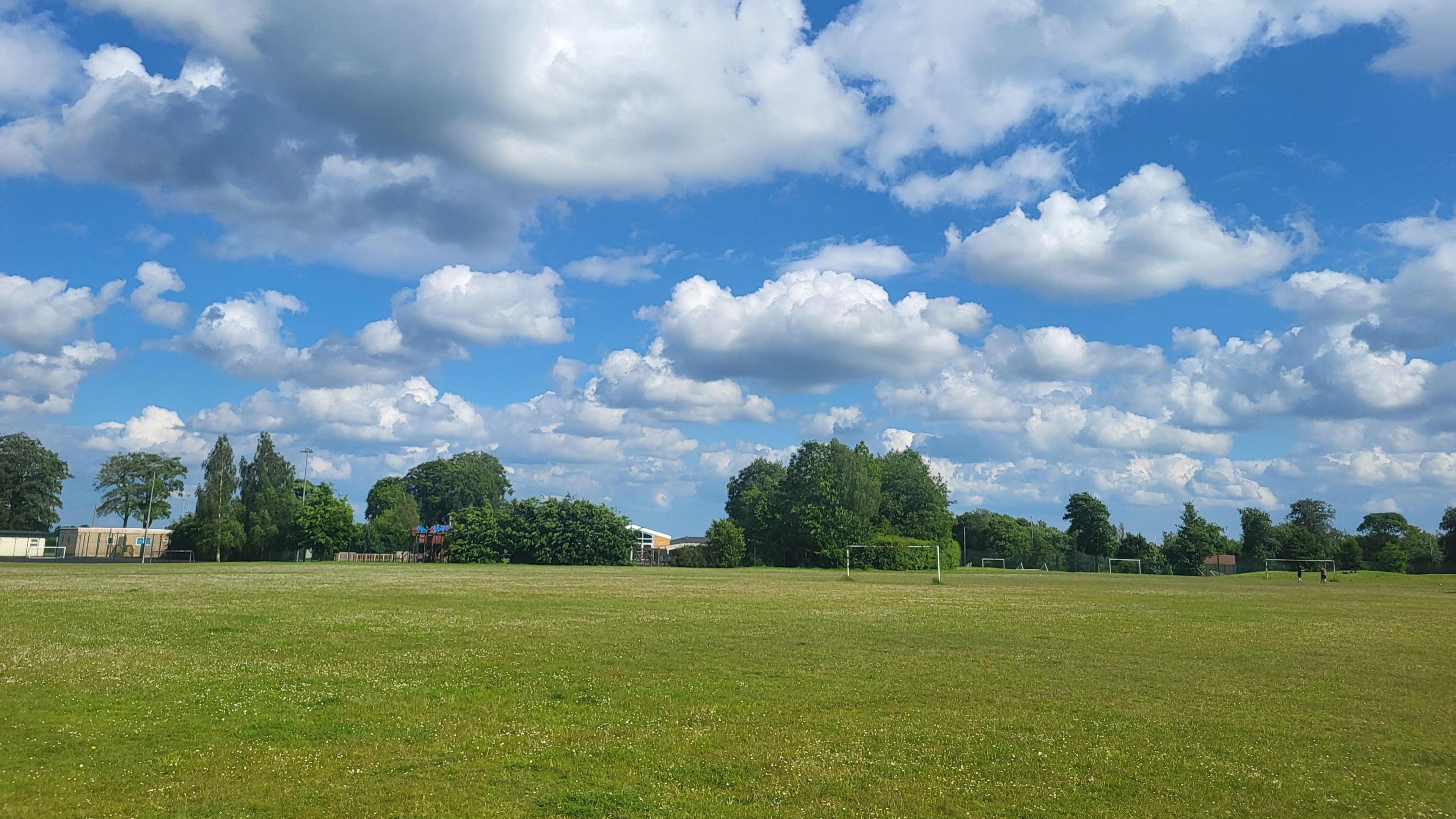 view of a green field with green trees in the distance.  There are sunny spells and fluffy white cumulus clouds in the sky