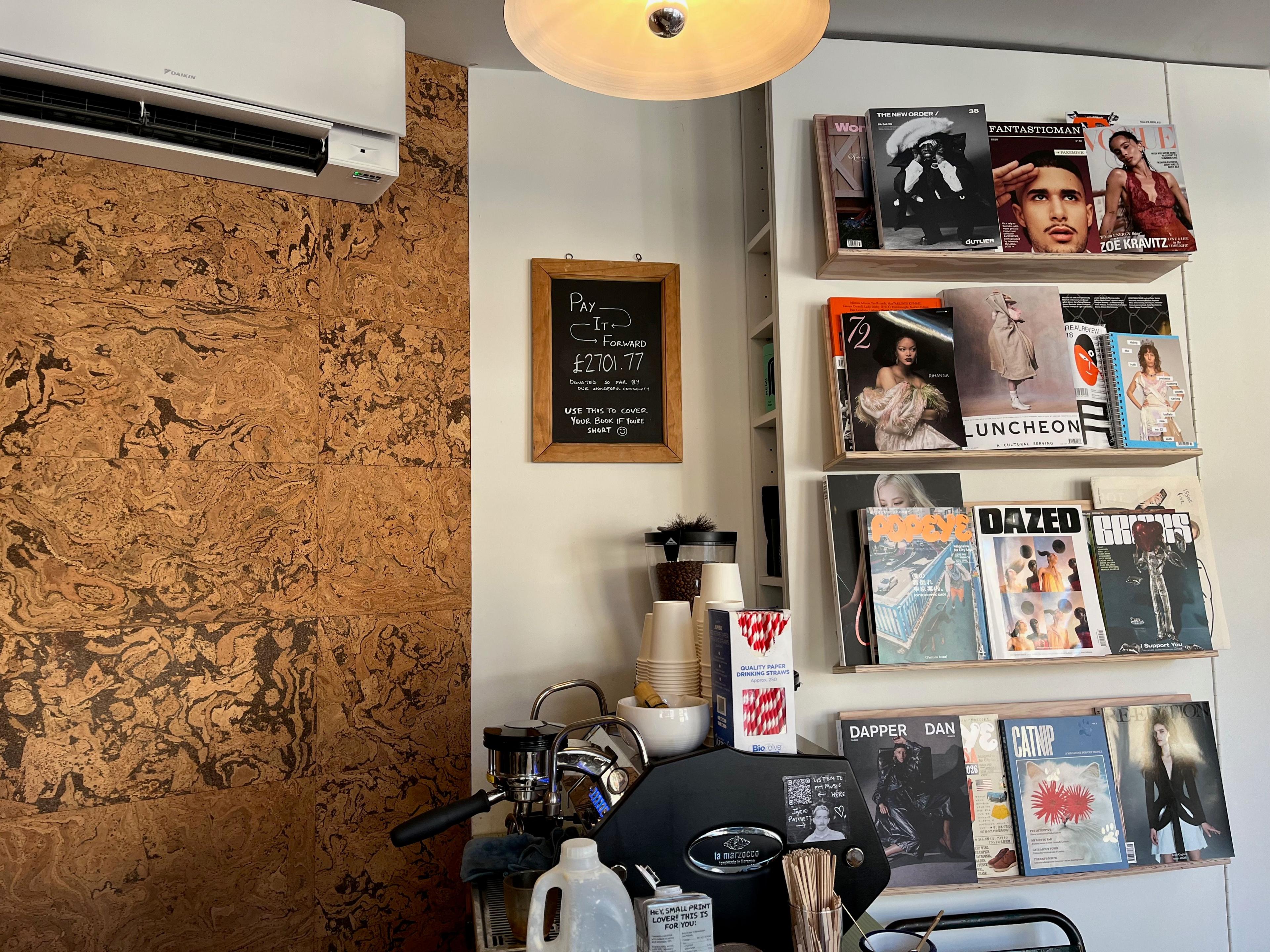 The interior of a book store with a display of magazines to the right, a coffee machine in the centre, and a black chalkboard sign that reads: Pay It Forward, £2701.77 donated so far by our wonderful community. Use this to cover your book if you're short. 