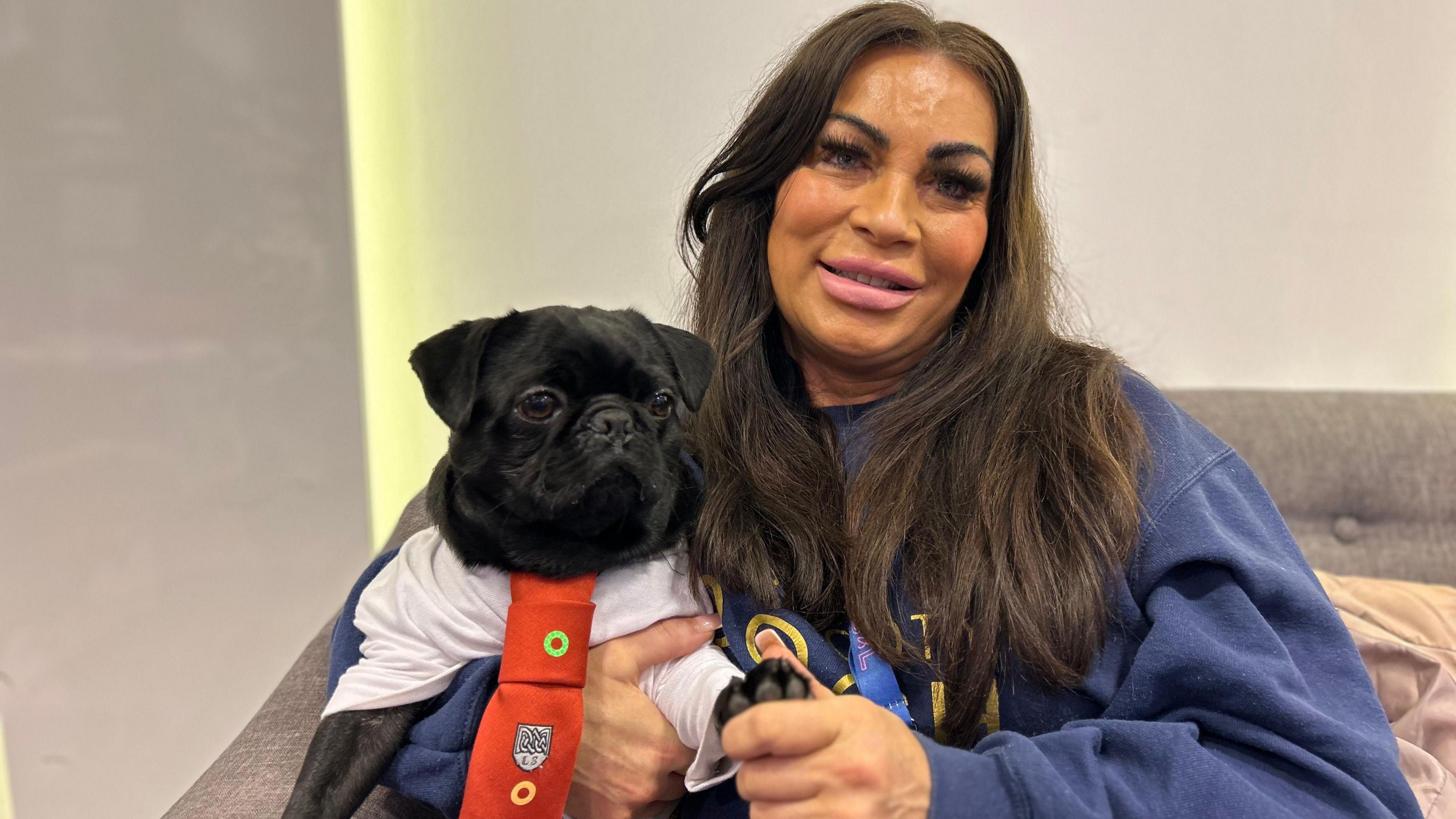 A woman in a navy blue sweatshirt with long black hair, posing with a black pug that is wearing a T-shirt and red tie with a school crest on it.  They are sitting on a grey sofa, with a grey wall behind.