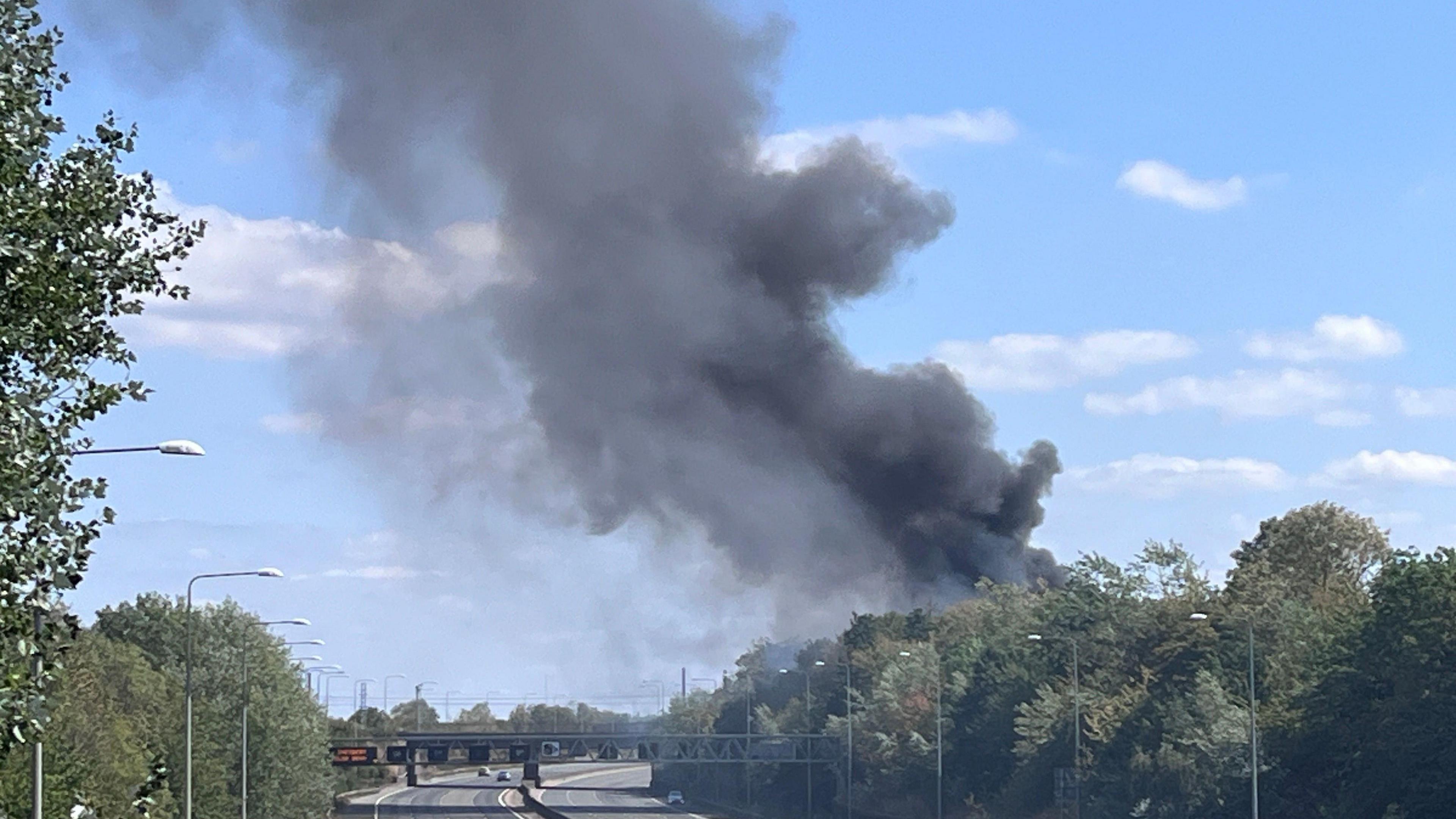 Smoke billowing across the M25, showing the road closed without any traffic on it. Trees are either side of the road.