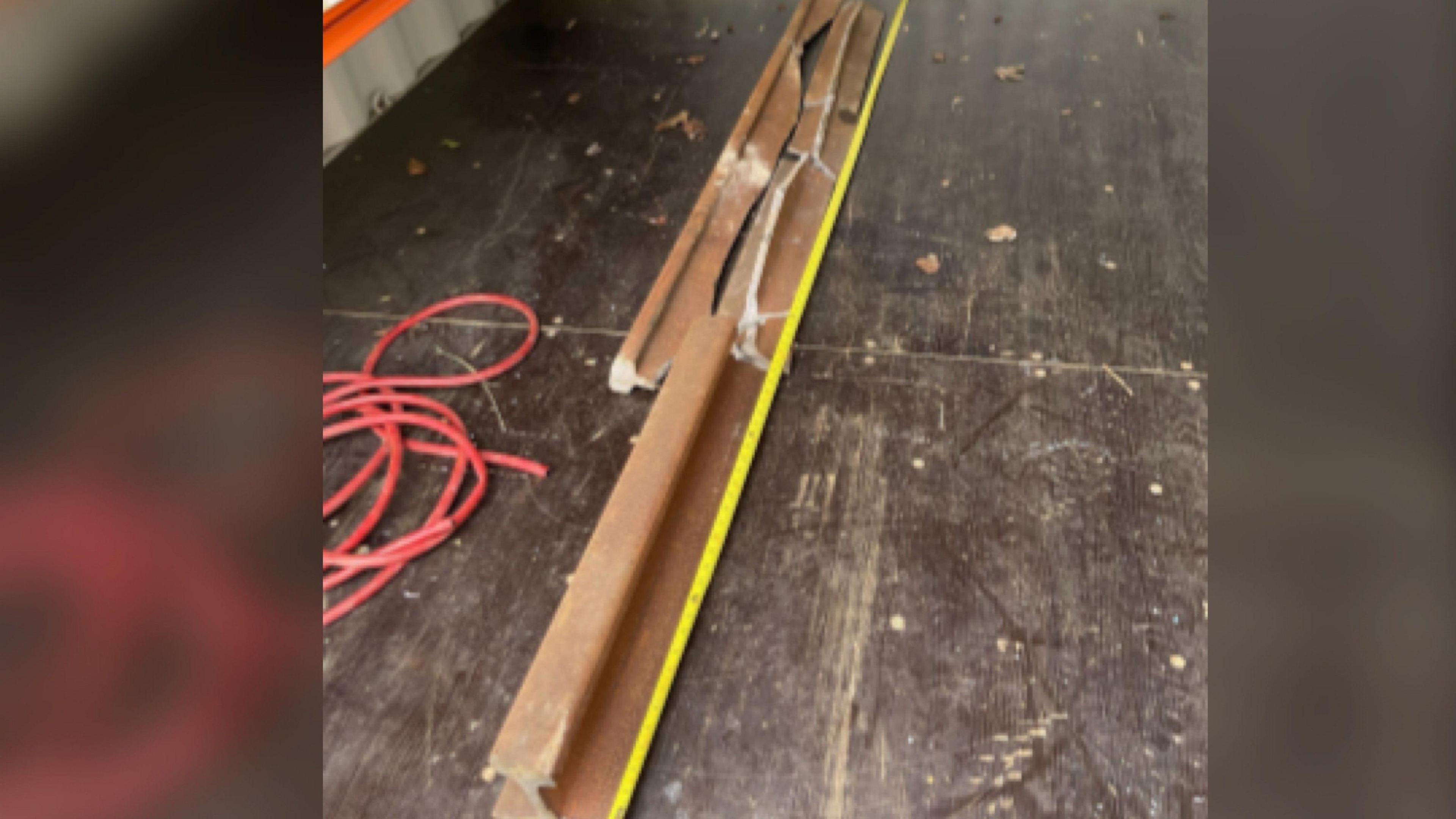 A rusty brown broken piece of rail track. It has a bright yellow stripe running alongside it. It has been photographed next to a red wire. 