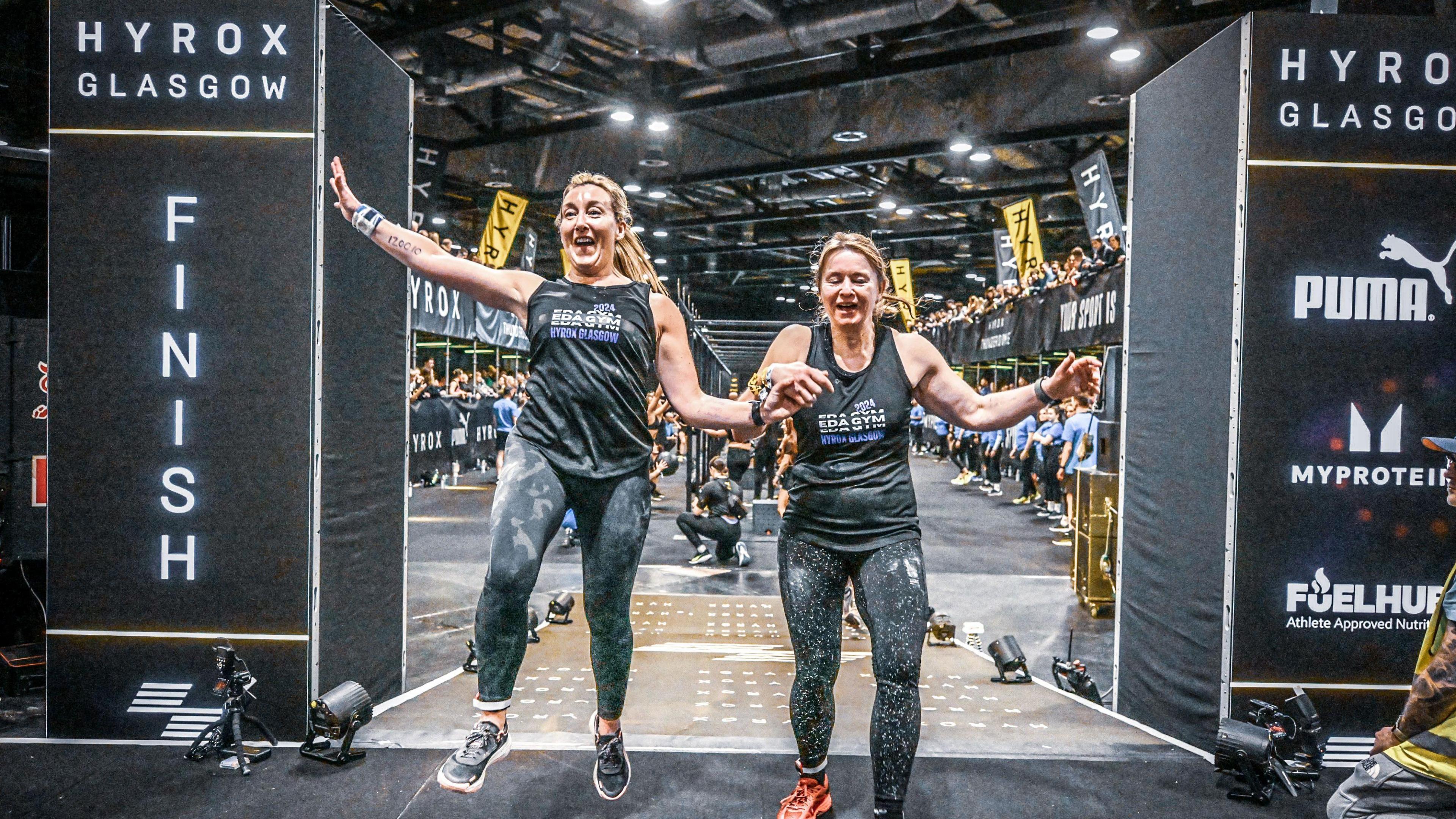 Leigh and her friend Wiki are smiling and holding hands as they cross the finish line at an indoor fitness race in Glasgow.