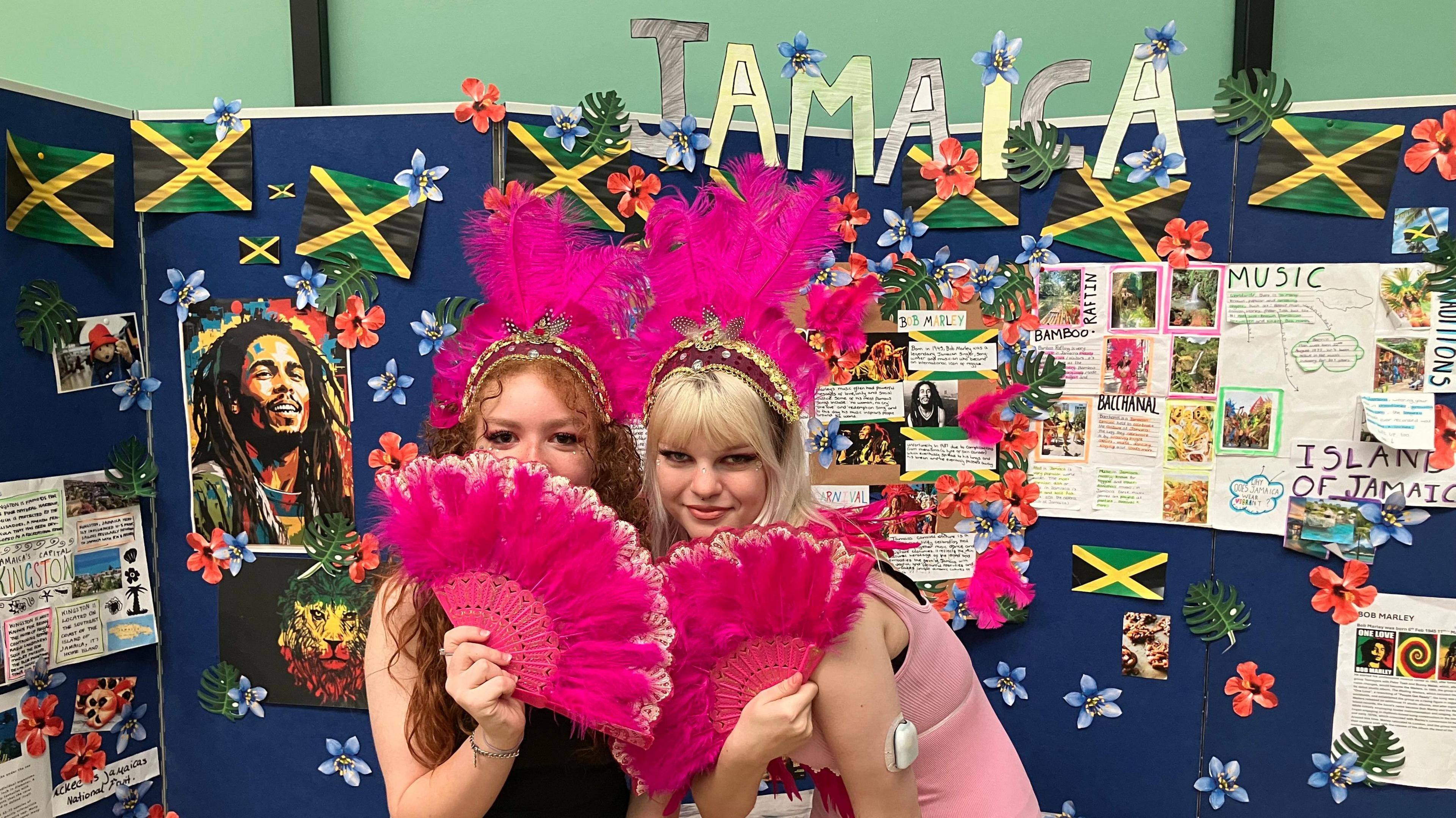 Roxen and Alex holding pink fans and dressed in carnival-style headpieces in front of the Jamaica stand