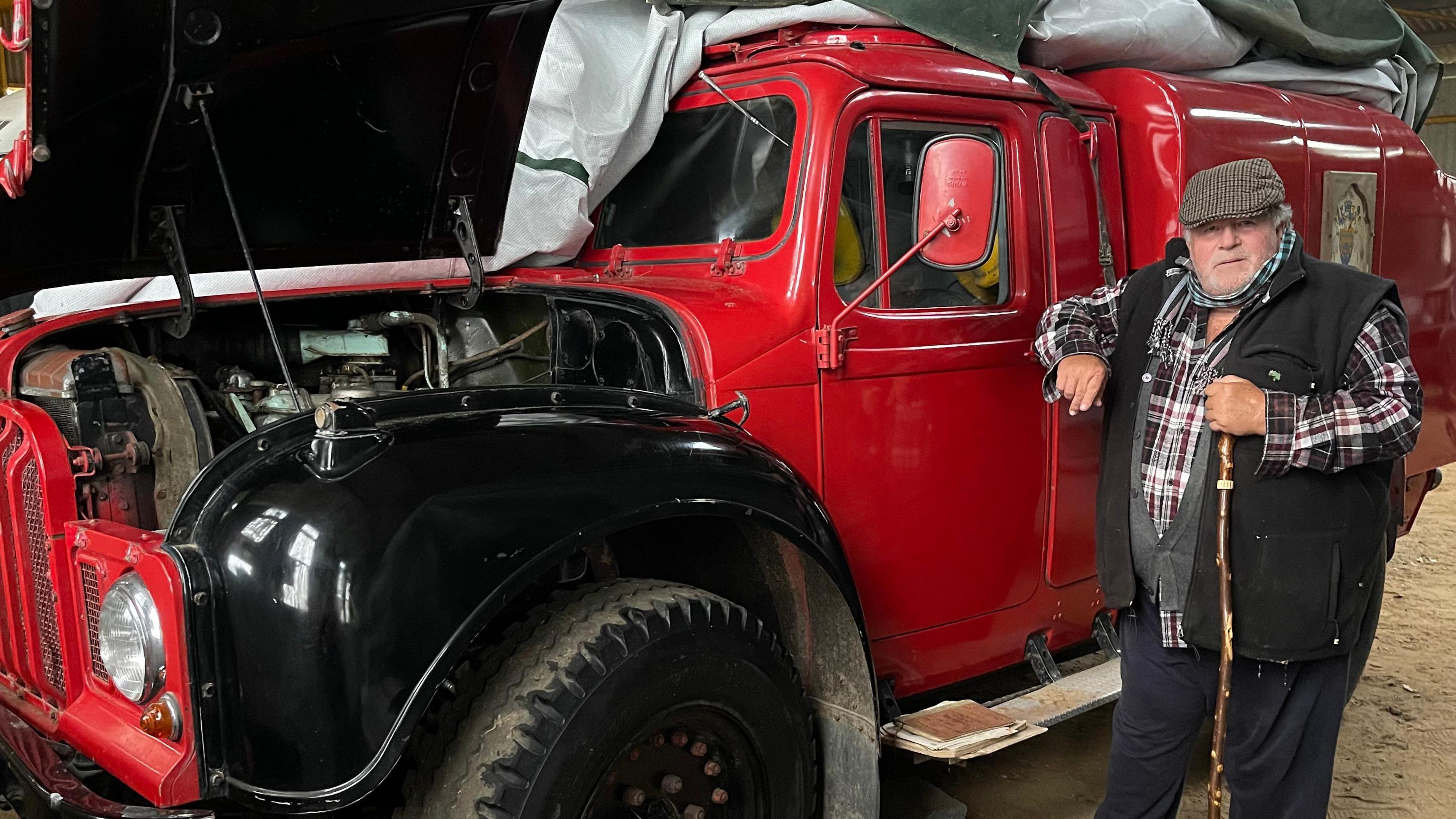 Richard Wallis wearing a checked shirt with a black fleece waistcoat with a flat cap on. He is standing next to the red and black rapid intervention vehicle, which has its bonnet partially open.