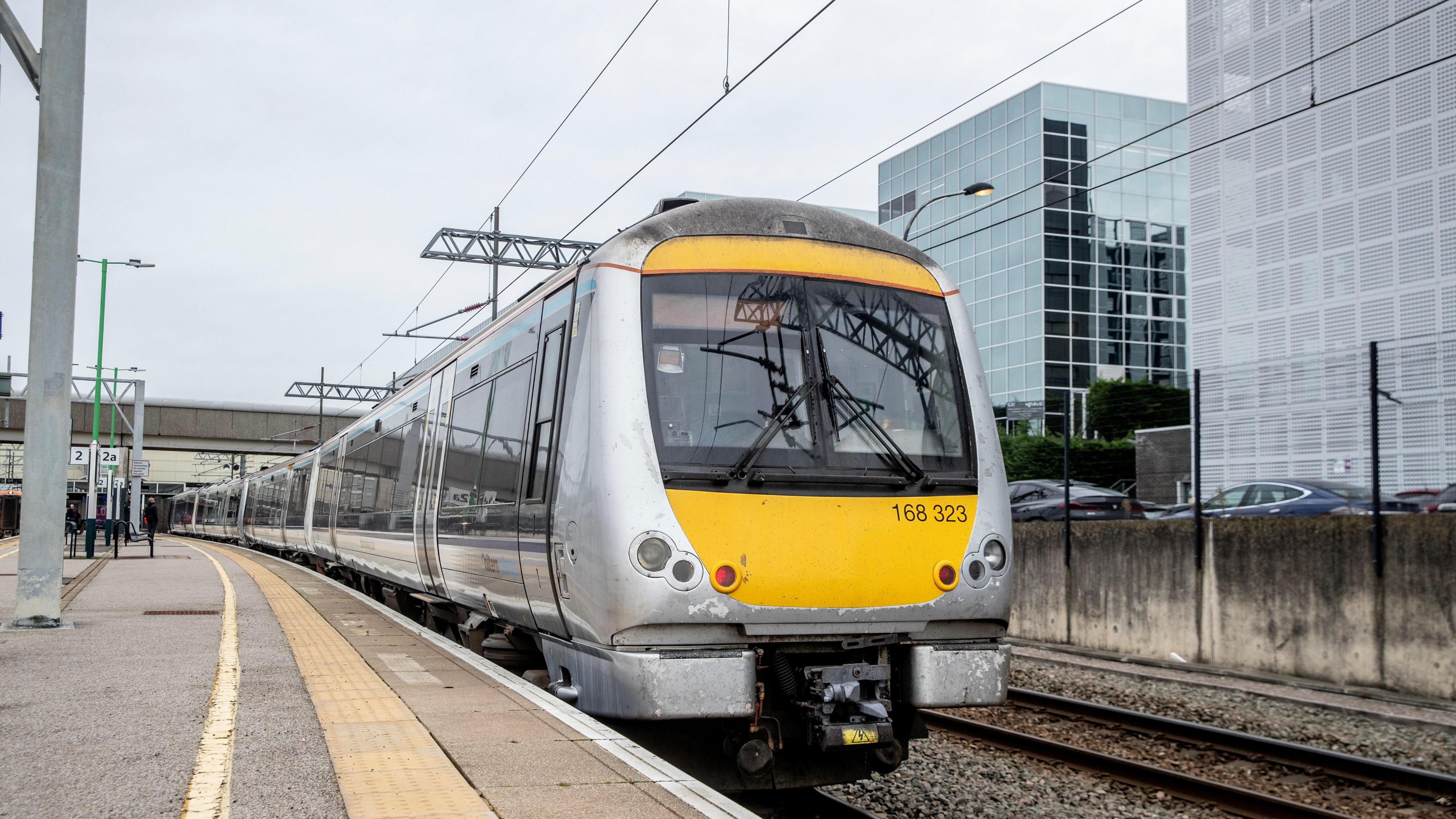 A silver and yellow train at Milton Keynes railway station, showing track buildings to the right and power lines.