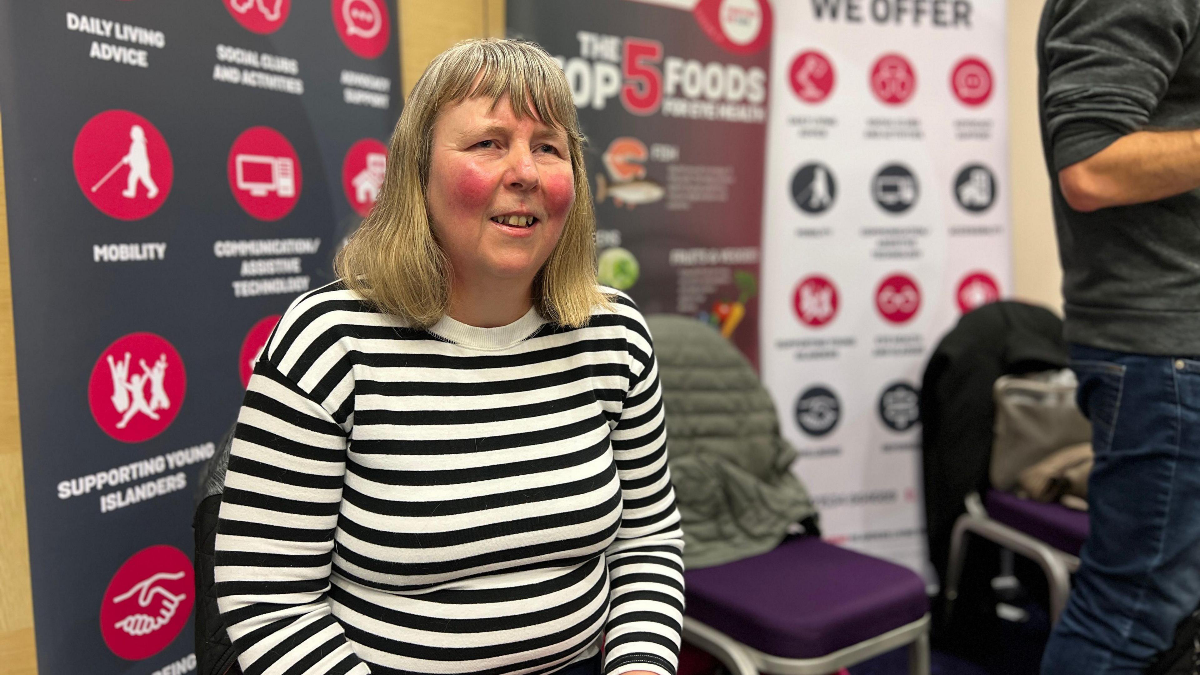 Jenny sits on a chair wearing a black and white stripped jumper and is sat in front of banners of the EYECAN charity. There are two chairs to her right which have coats and bags on them. Jenny has light-coloured hair and it comes down to her shoulders.