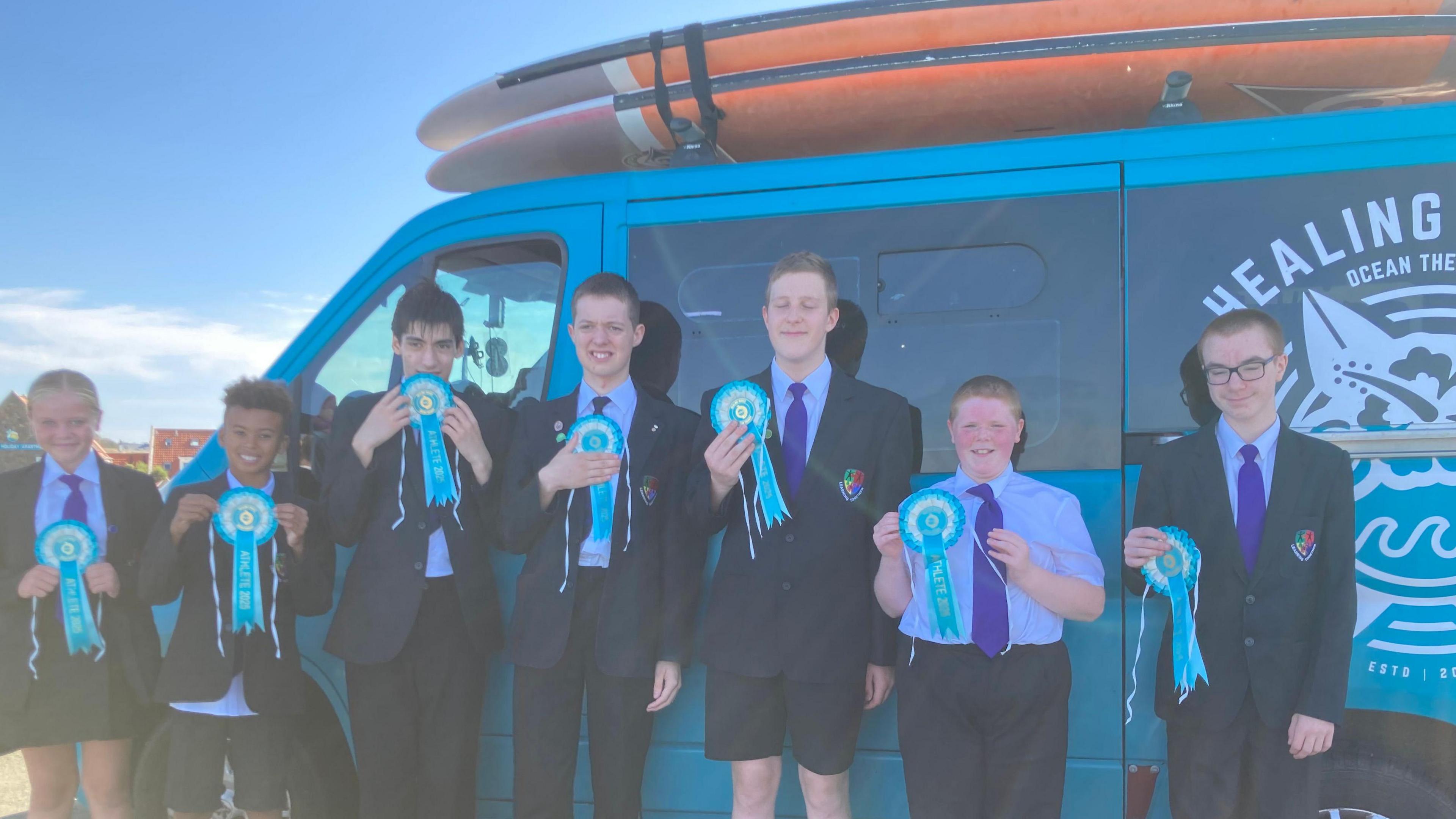 A group of school children wearing navy suits with a paler blue shirt and bright blue tie stand in front of a van with a surf board on top. they are all smiling and holding up turquoise rosettes.