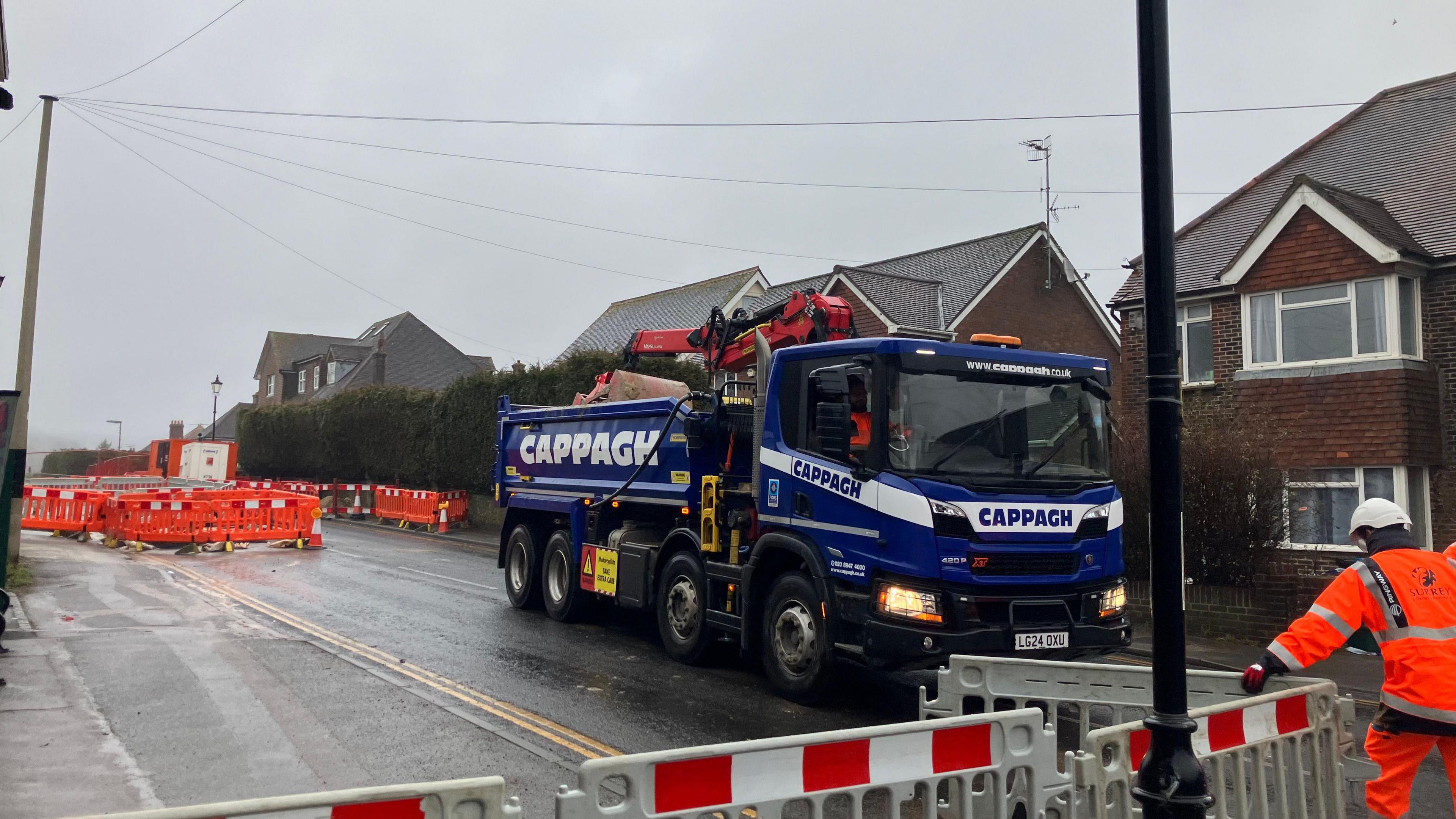 A blue tipper lorry loaded with sand is driven away from the site of two sinkholes that have appeared in Godstone, Surrey. Two men wearing hi vis are opening a road barrier to allow the lorry to pass through.