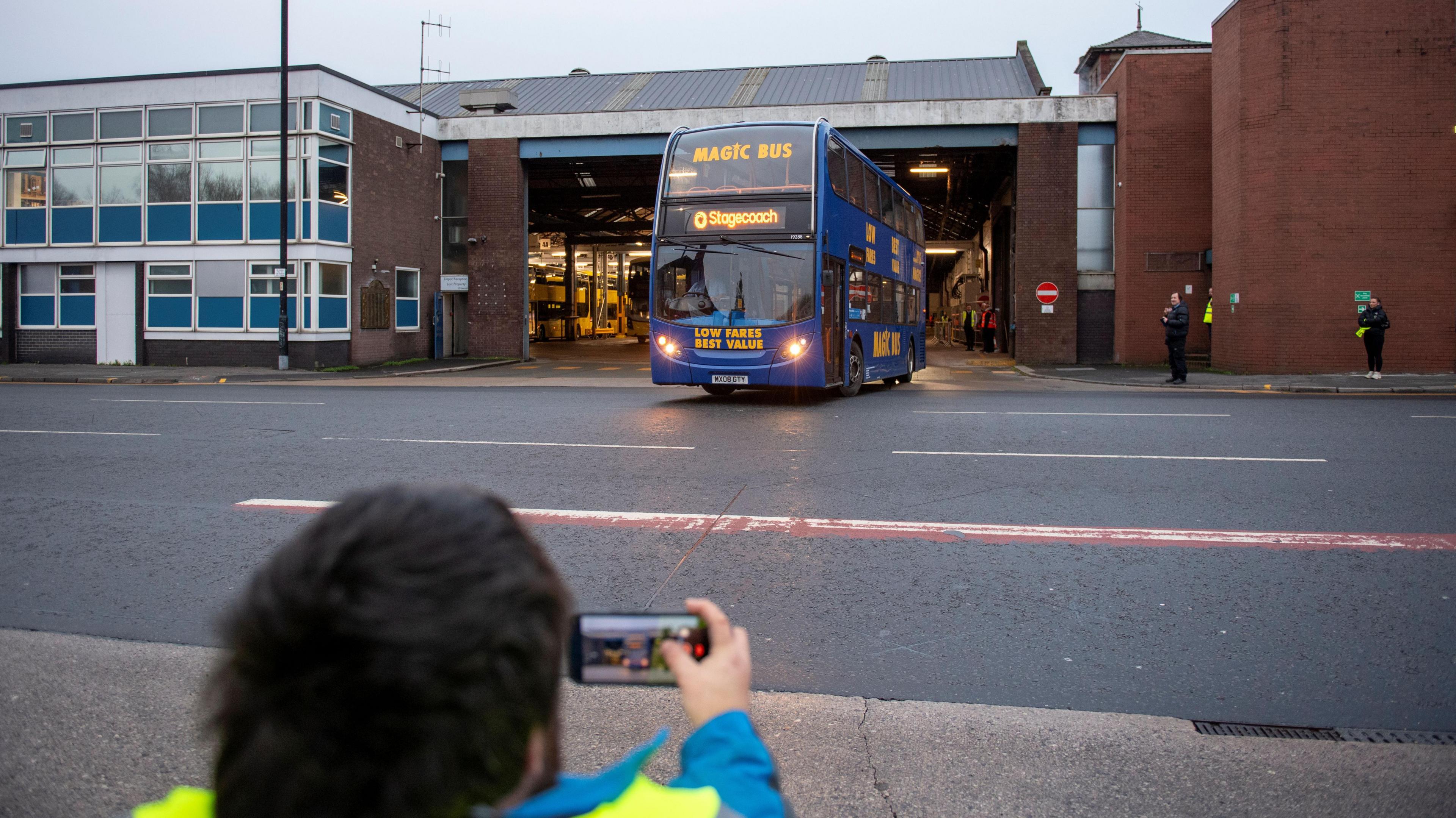 A man films a Magic Bus drawing out of a garage through his smartphone. 