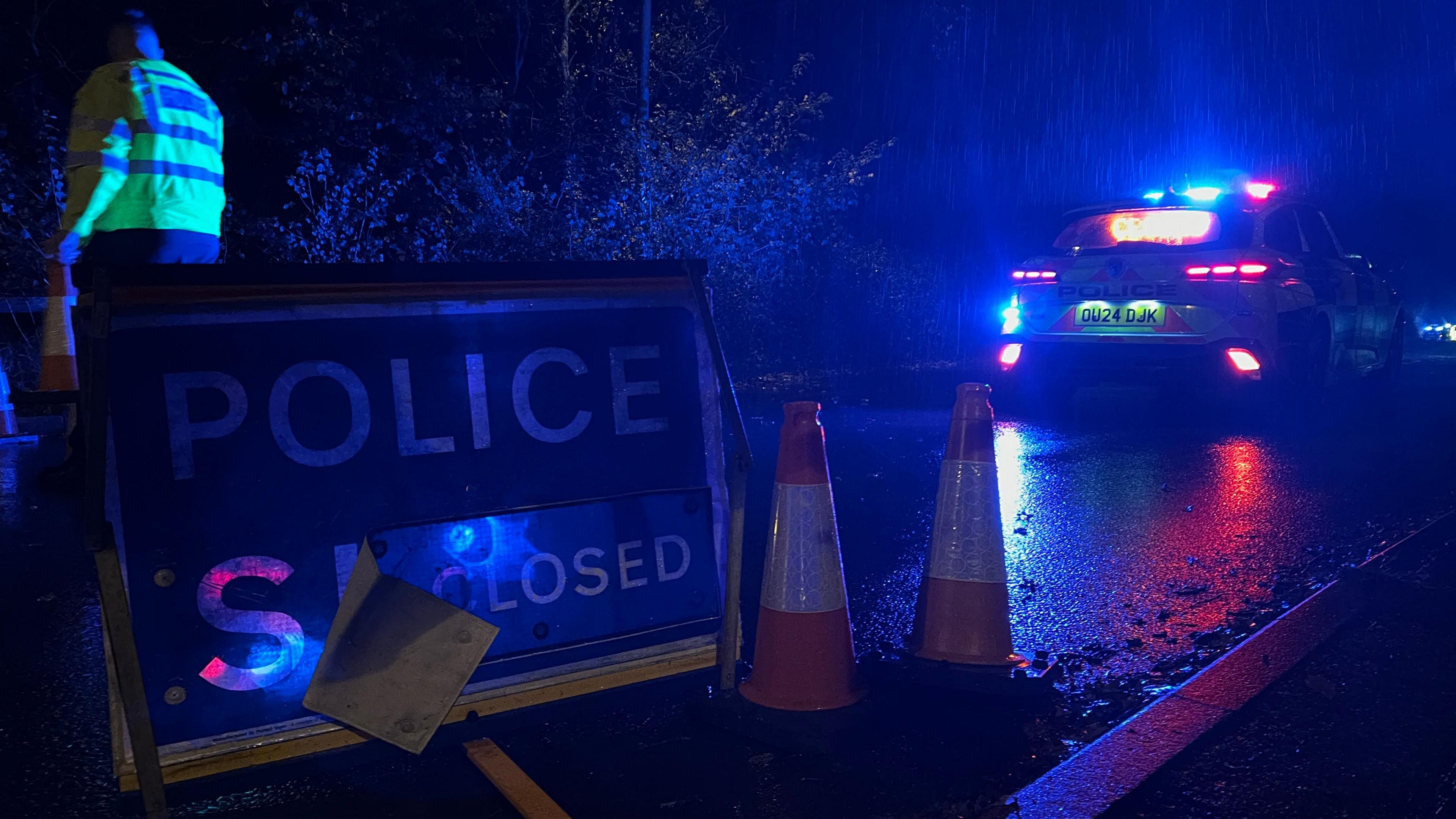 A blue sign advising people that the road has been closed by the police. It is next to traffic cones and a police car that has its lights on. The picture, which is an archive image, has been taken in the evening.