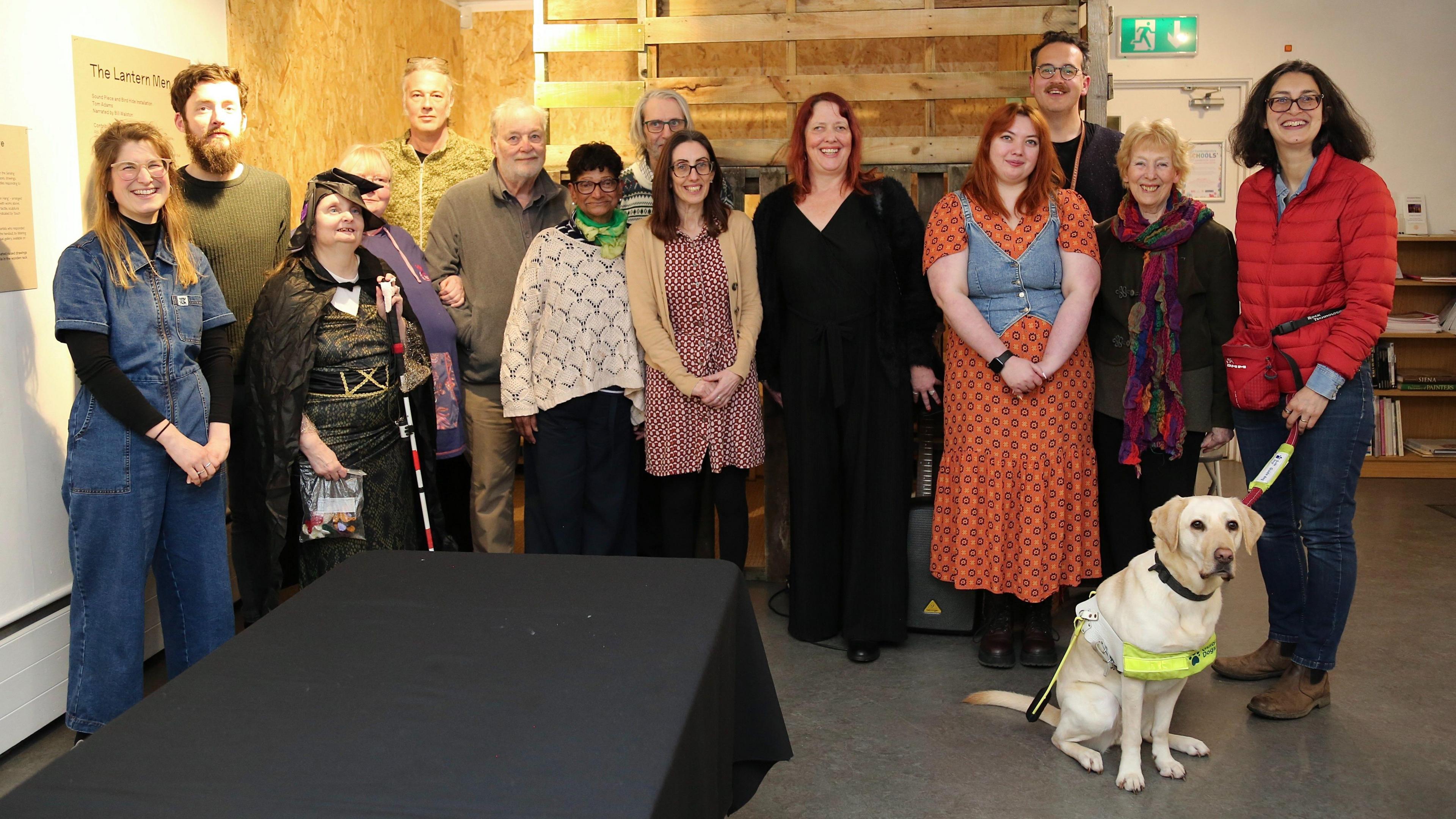 A group of people, standing looking at the camera, at a listening party at Babylon Arts. It shows a woman with a guide dog, a woman, dressed in a costume with a stick, and a mixture of men and women. 