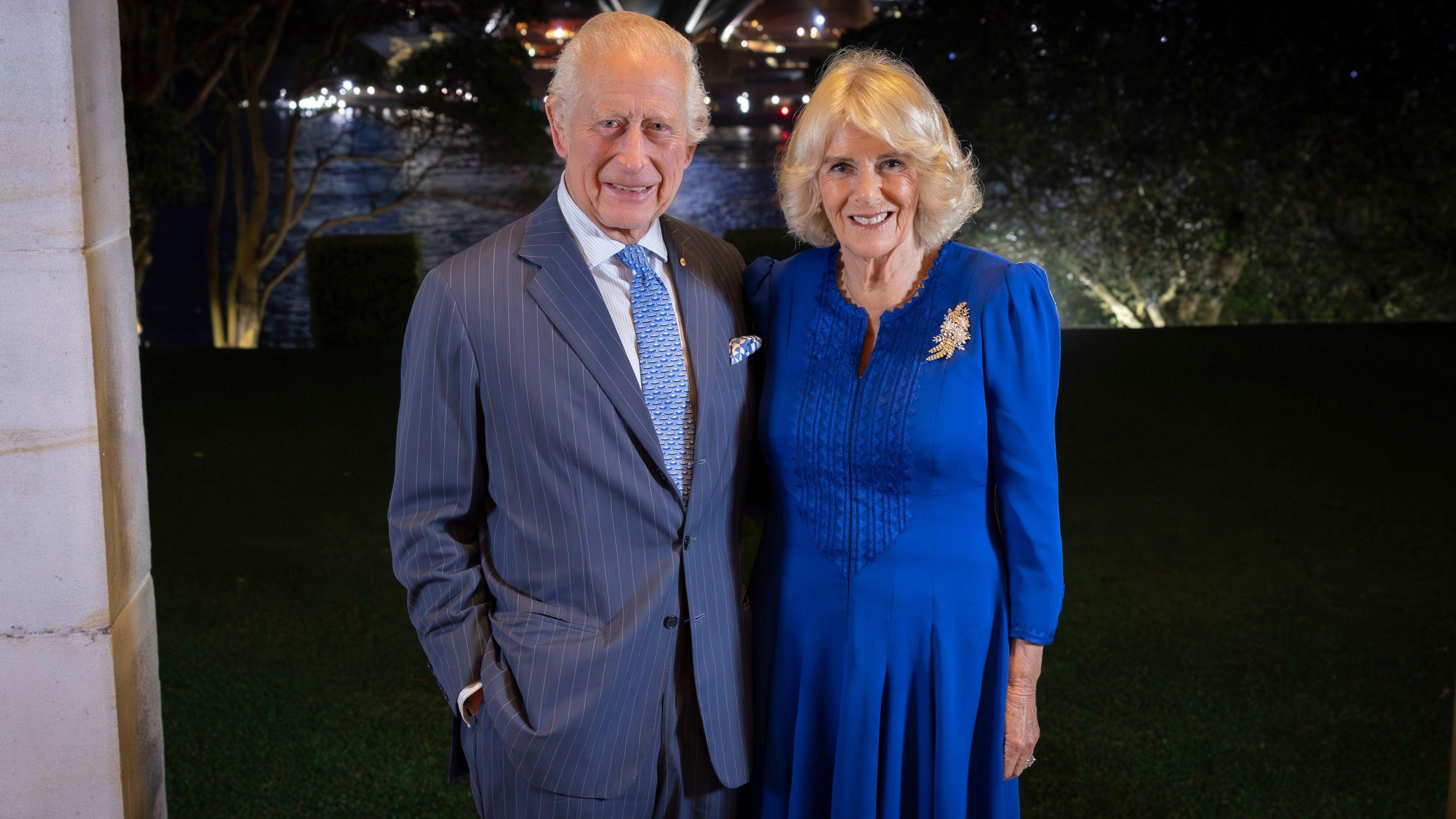 King Charles and Queen Camilla pose for the camera with the Sydney Opera House in the background at night. The King is wearing a grey striped suit with a light blue tie and the Queen is wearing a blue dress with a broach.