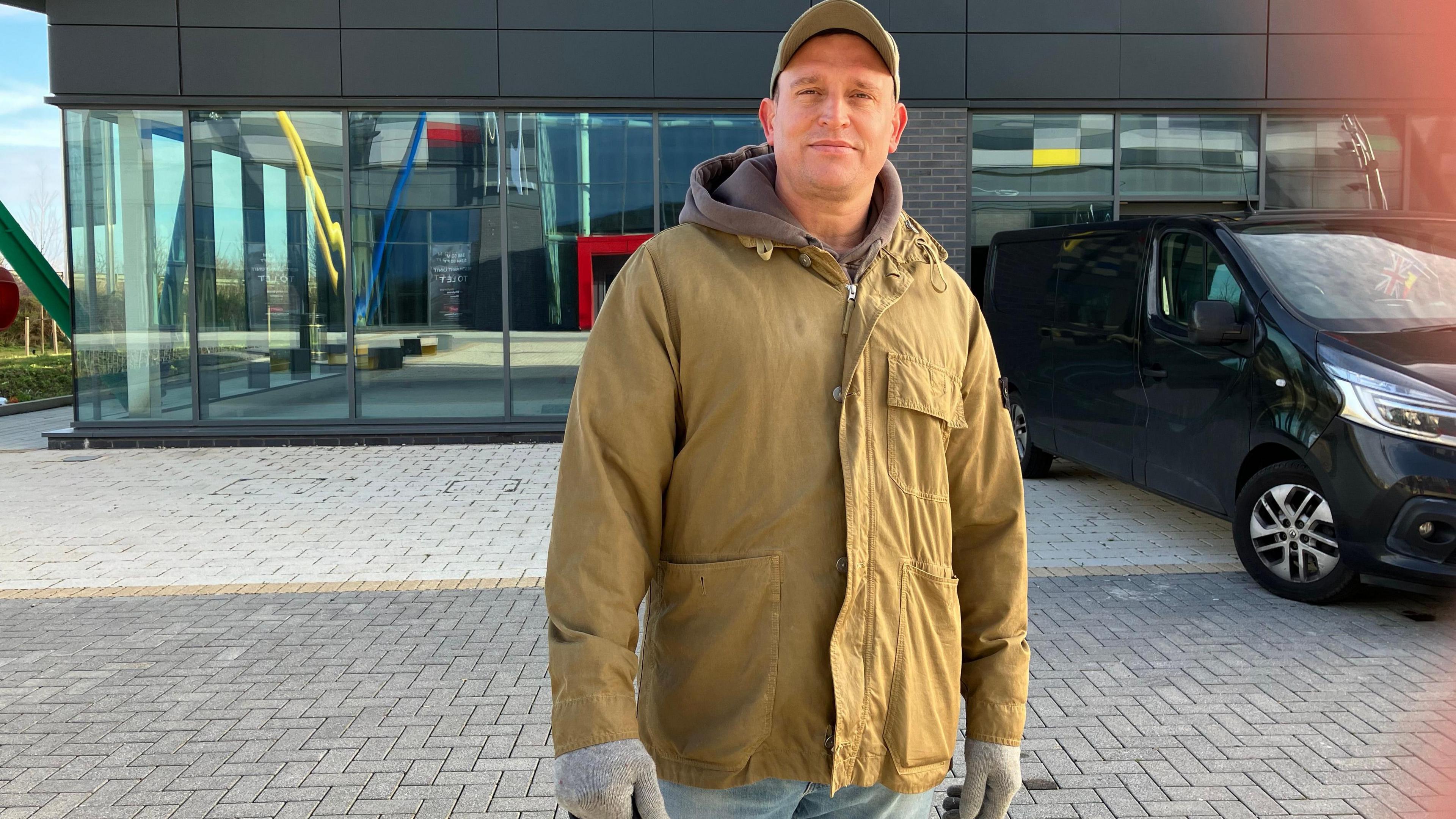 Glenn Matthews in a baseball cap and a light brown winter coat standing in front of a modern-looking building at the leisure park. He has a dog on a lead. The dog is not in shot. There is a black car in front of the building.