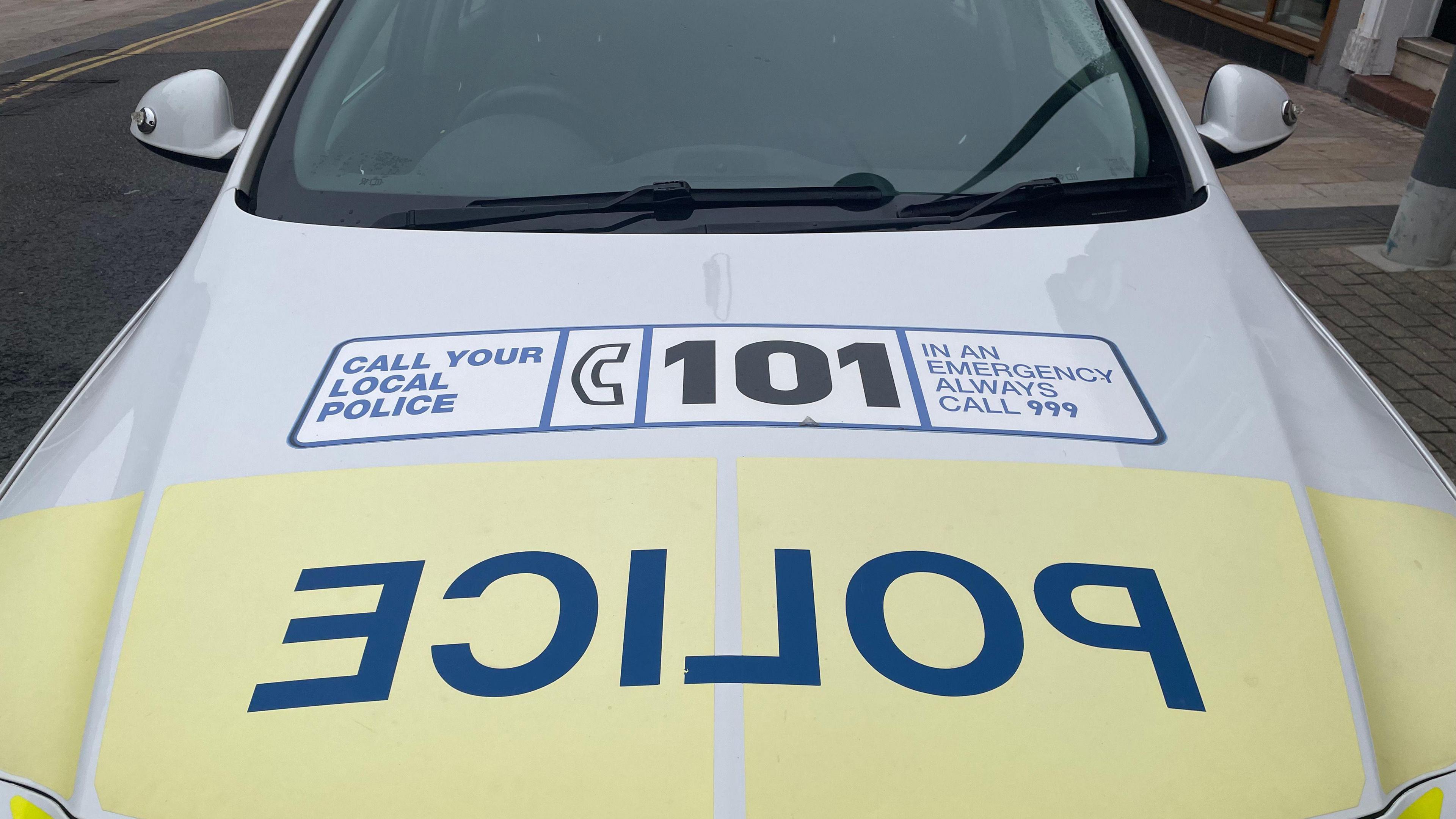 The bonnet of a yellow and white police car with the word police written backwards in large blue letters and a label telling people to call 101