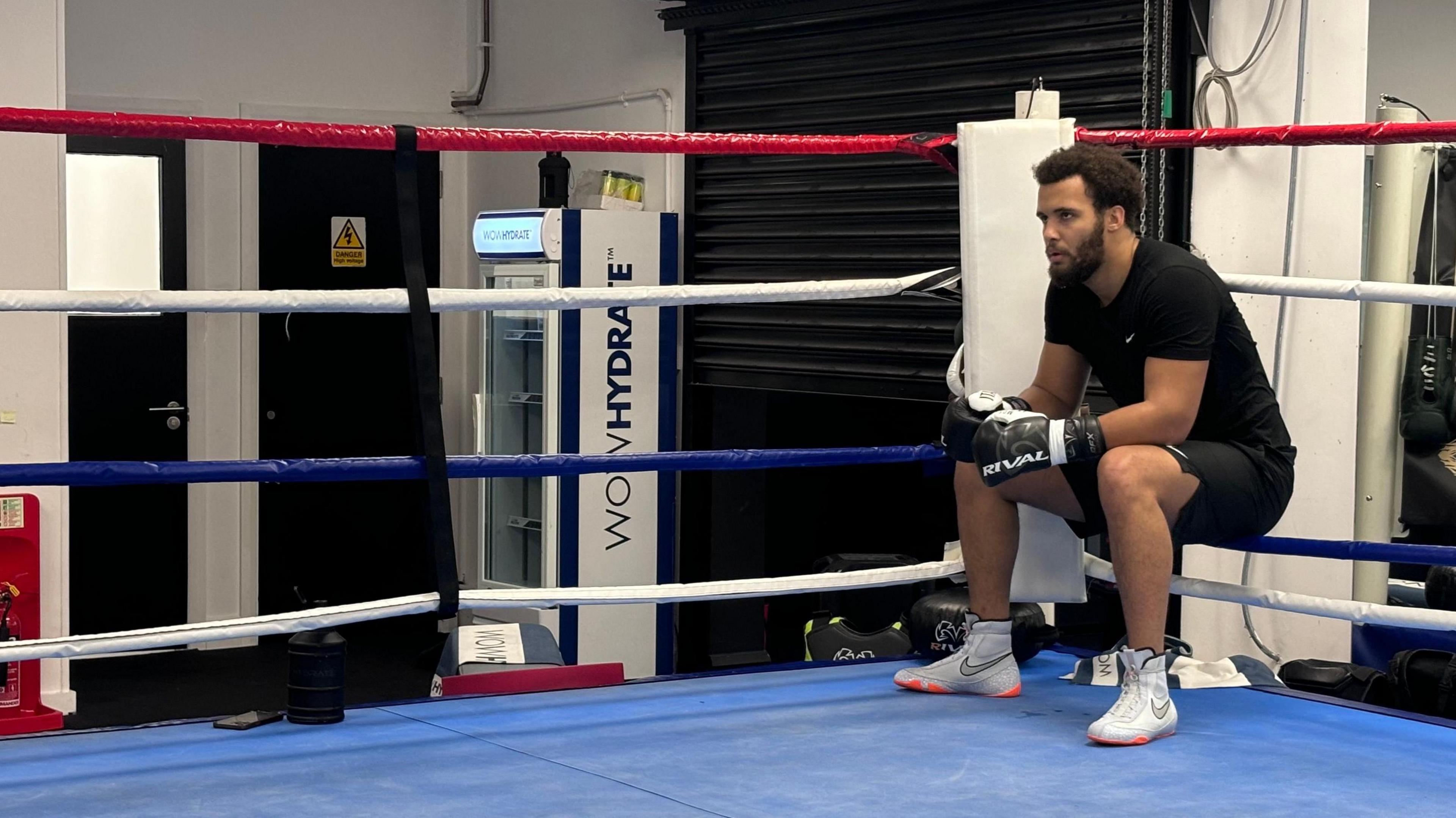 Moses Itauma at a gym in Essex where he is training for his next fight. He is wearing a black top and shorts, and black boxing gloves. He is sat on the bottom of four ropes inside a boxing ring. 