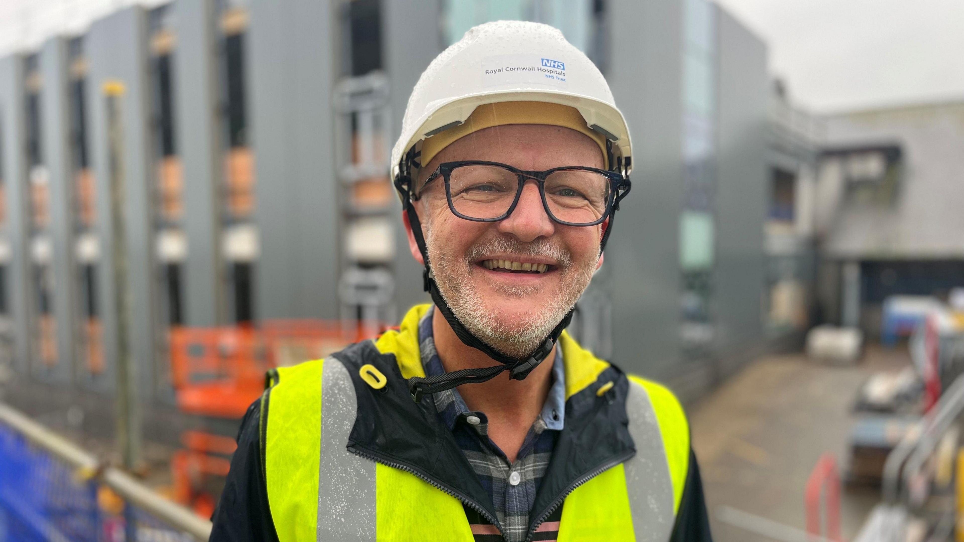 Paul Sylvester is smiling at the camera with the new building behind him. He is wearing a hard hat and a high-viz waistcoat.