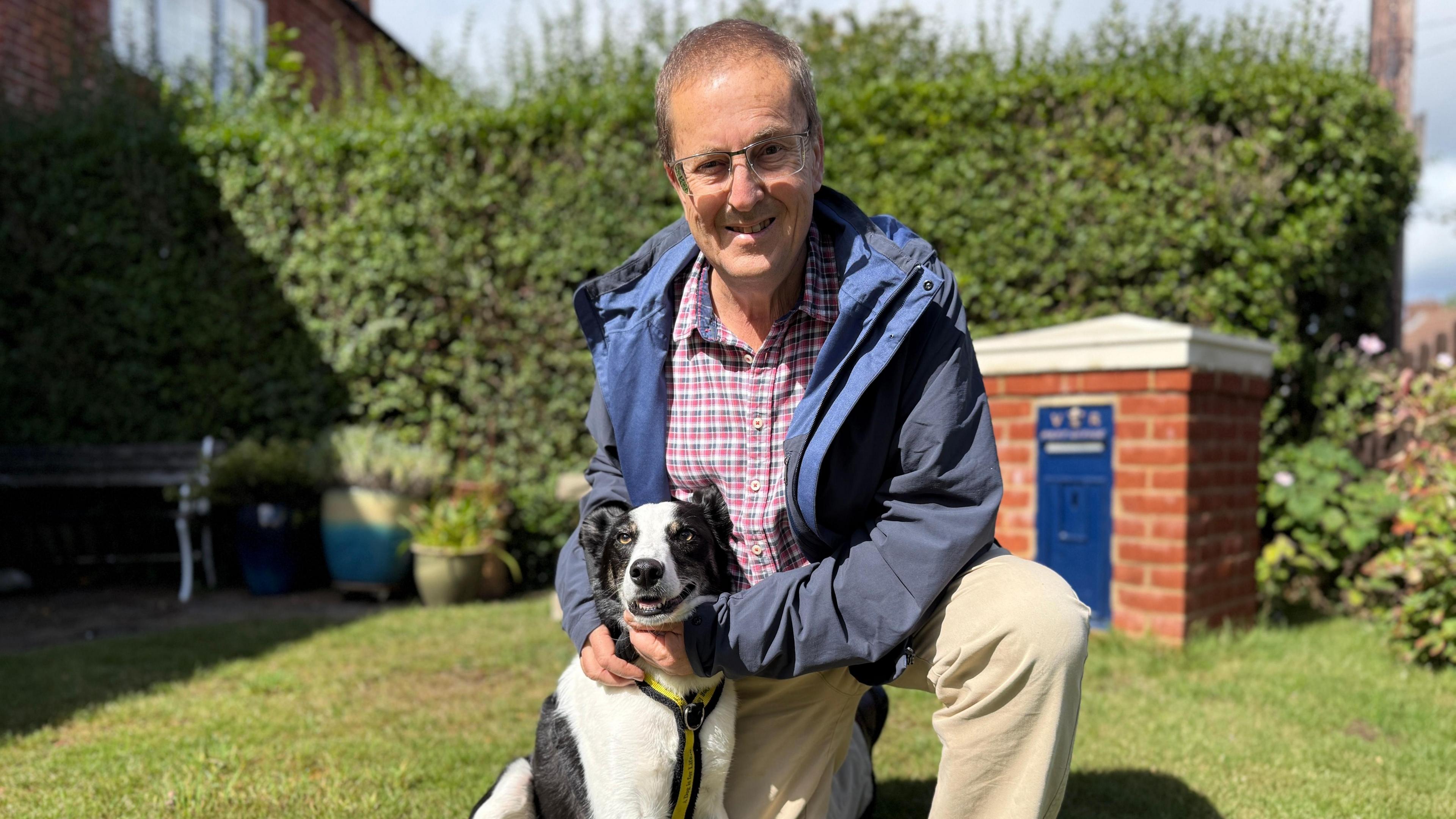The image shows David crouched down on one knee. Next to him is his black and white dog Juke. He and his dog are in a garden. Behind them is a green bush. In front of the bush are a few plant pots and a garden bench. On the right, there is an orange brick structure with a white peaked roof. On the front of the structure, there is a blue panel that looks like a mini postbox. David is wearing a red and blue shirt, blue waterproof jacket and beige trousers. He is wearing glasses and smiling at the camera. His dog Juke is also looking at the camera. Juke has a yellow and black harness.