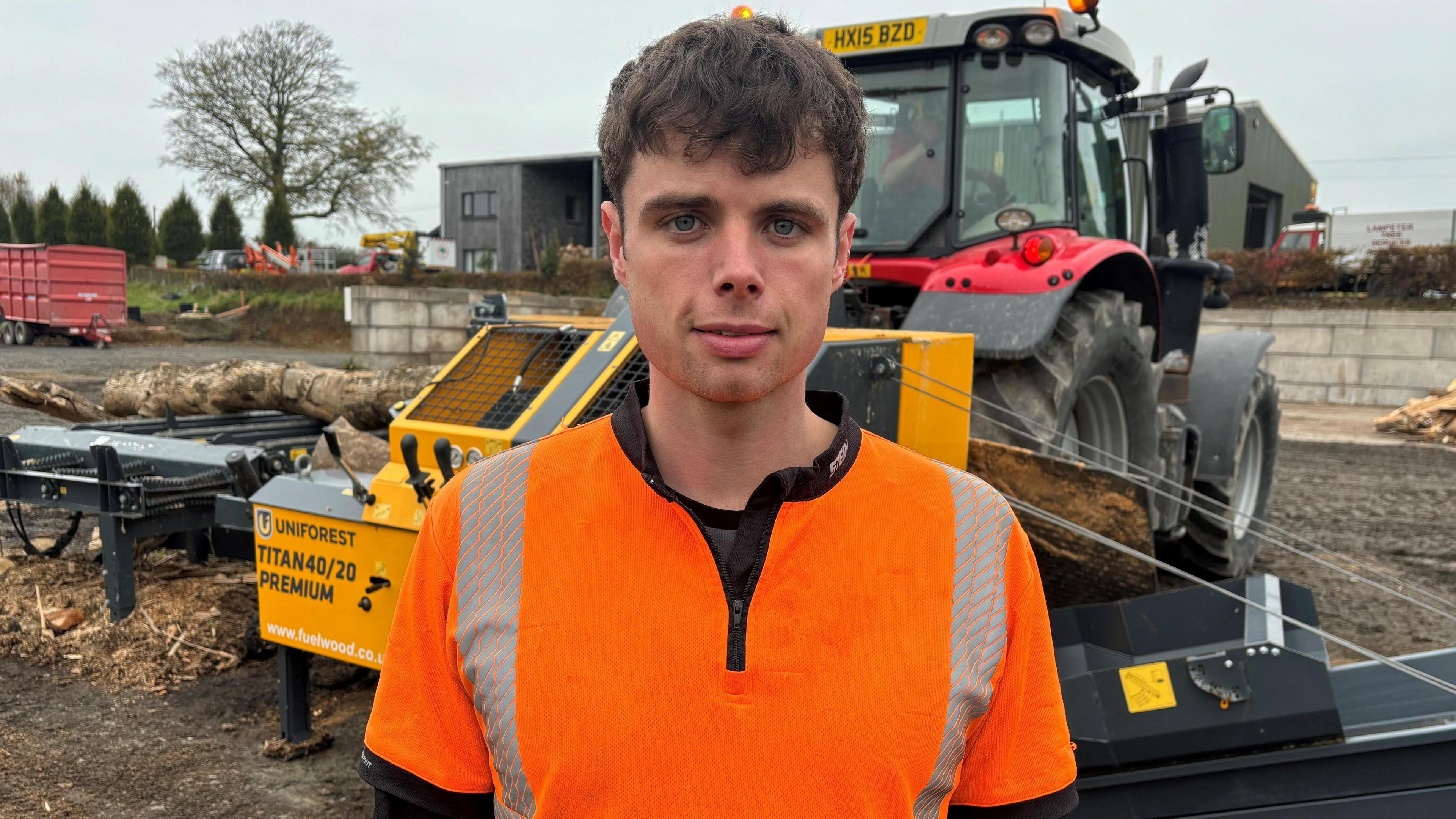 Tomos Williams yn sefyll mewn hi-vis oren o flaen cloddiwr