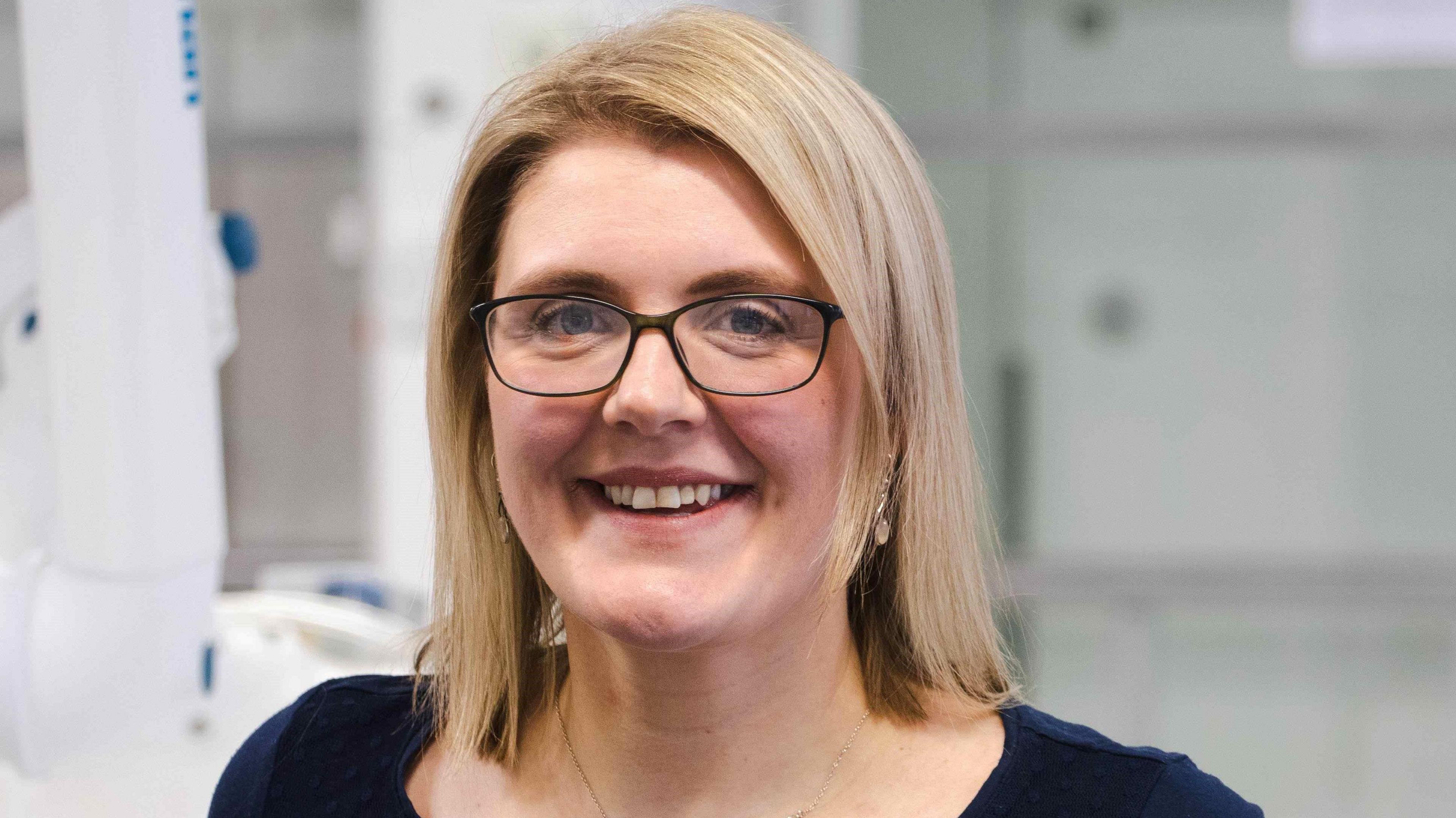 A blonde woman with glasses smiling at the camera. It is a head and shoulders shot of her.
