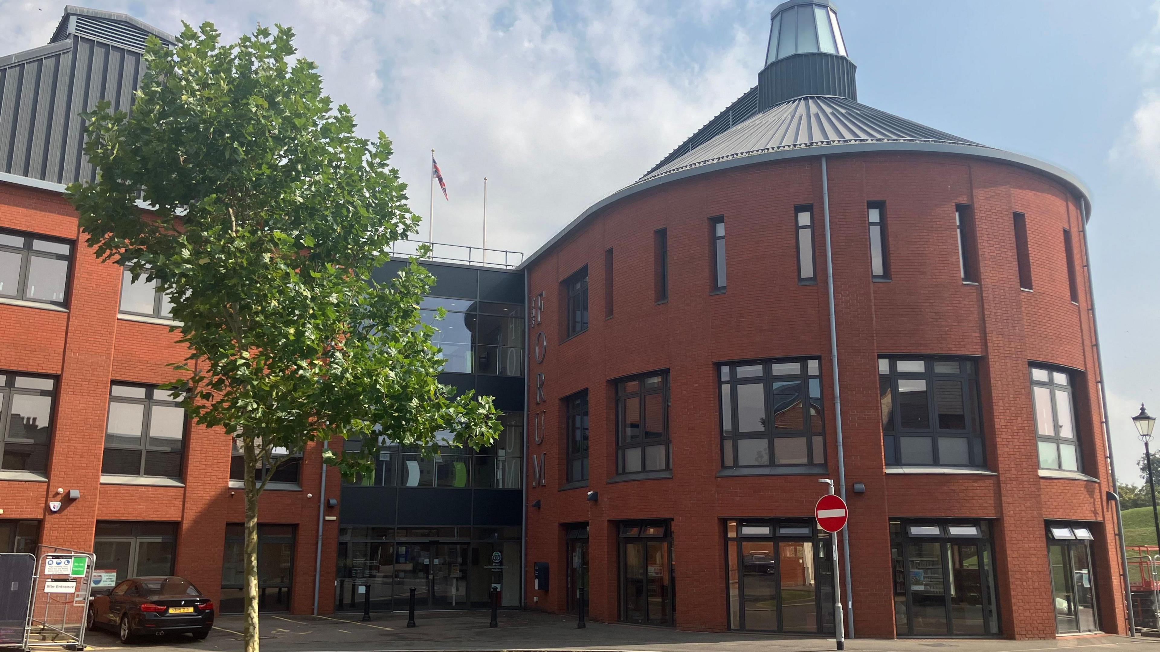 A council building made out of red brick. The building is curved and has grey-framed windows. There is a tree outside and a no entry sign. A car is parked outside the building. 