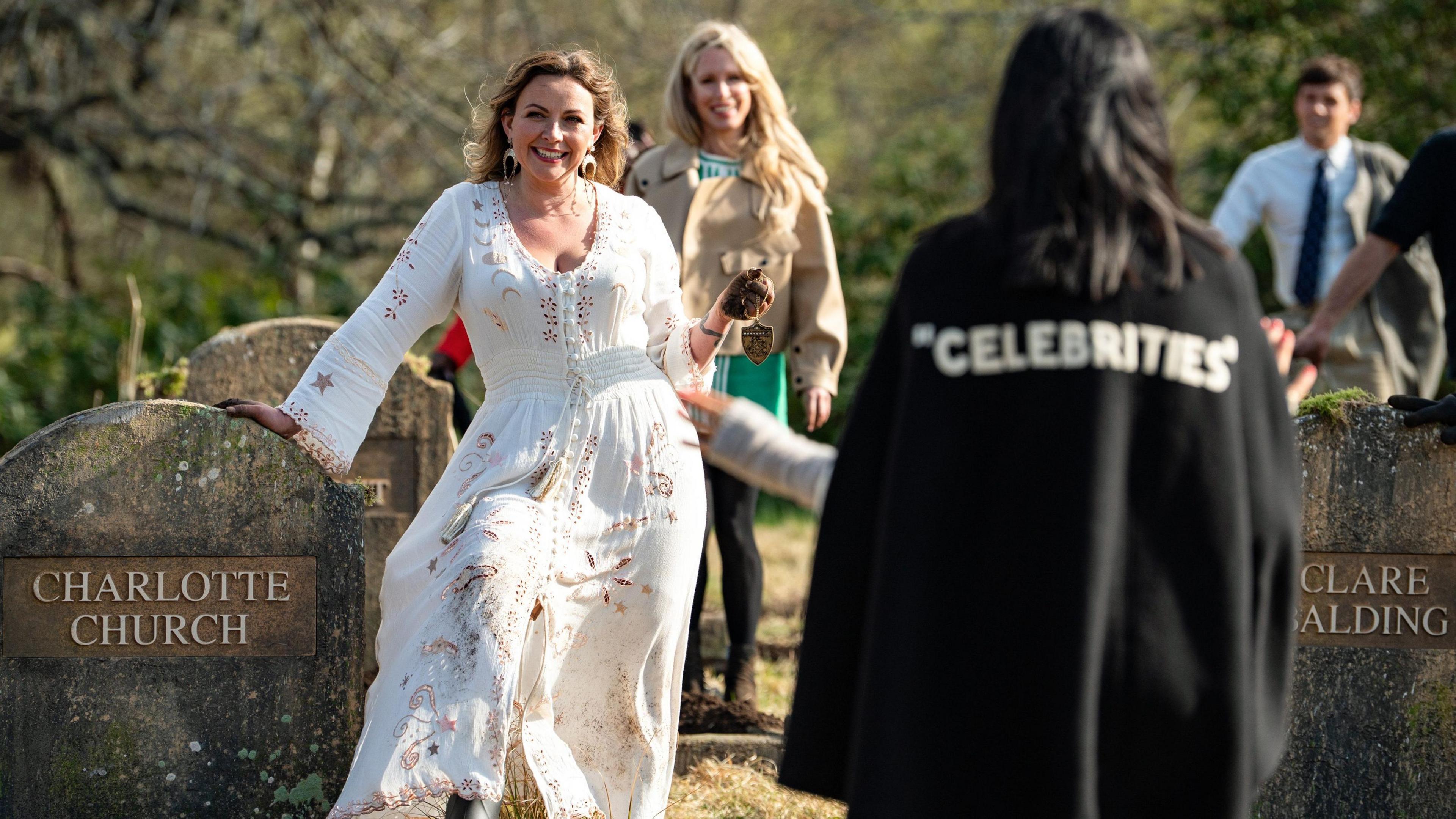 Claudia Winkleman wearing a black cloak with the word "celebrities" as the cvelebrities arrive at a graveyard with gravestones showing their names