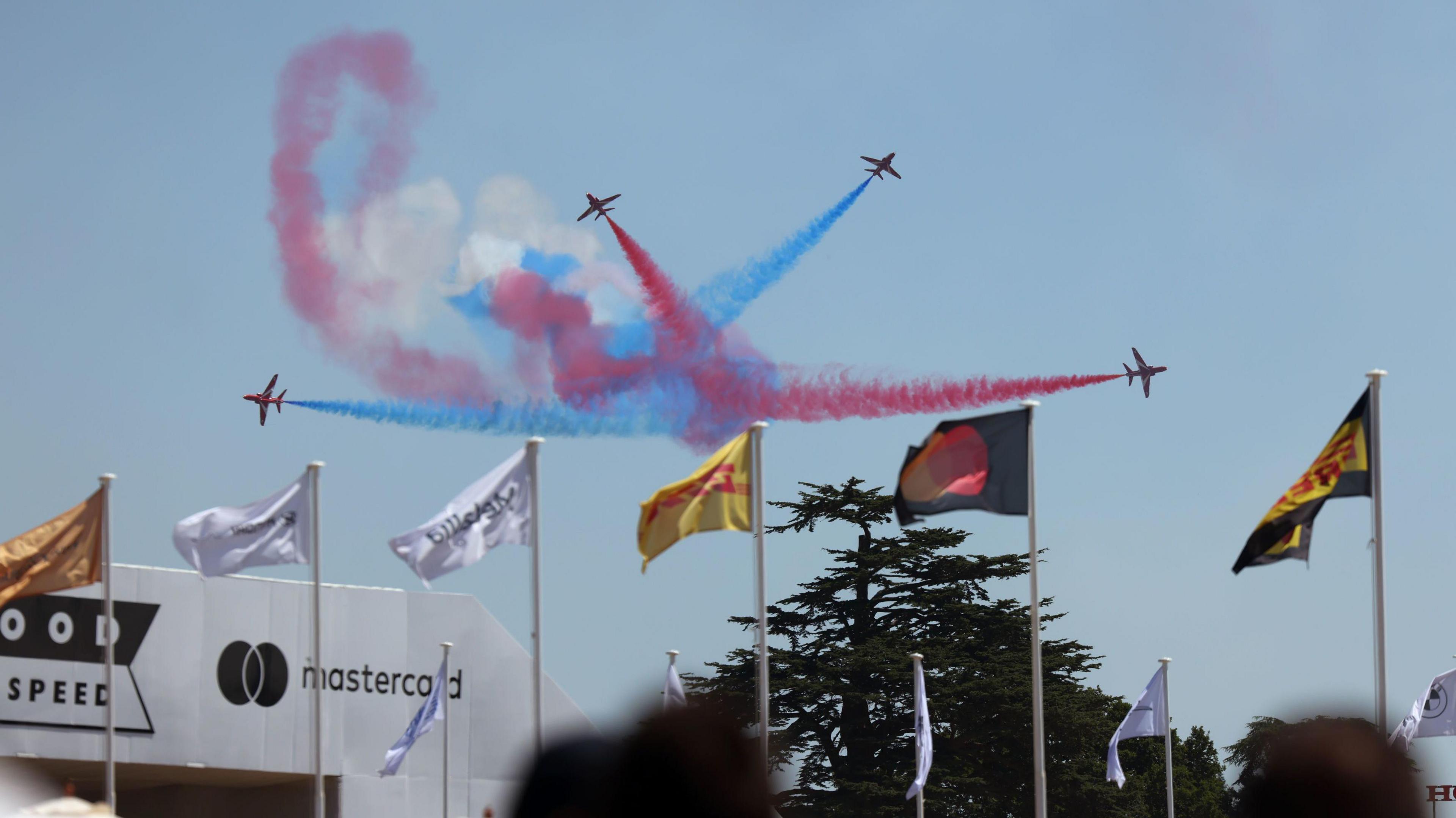 Four red arrow planes fly in different directions, trailing red, blue and white vapour behind them. They are flying against the backdrop of a blue sky, and there are six flags in the foreground, and a tree in the background. 