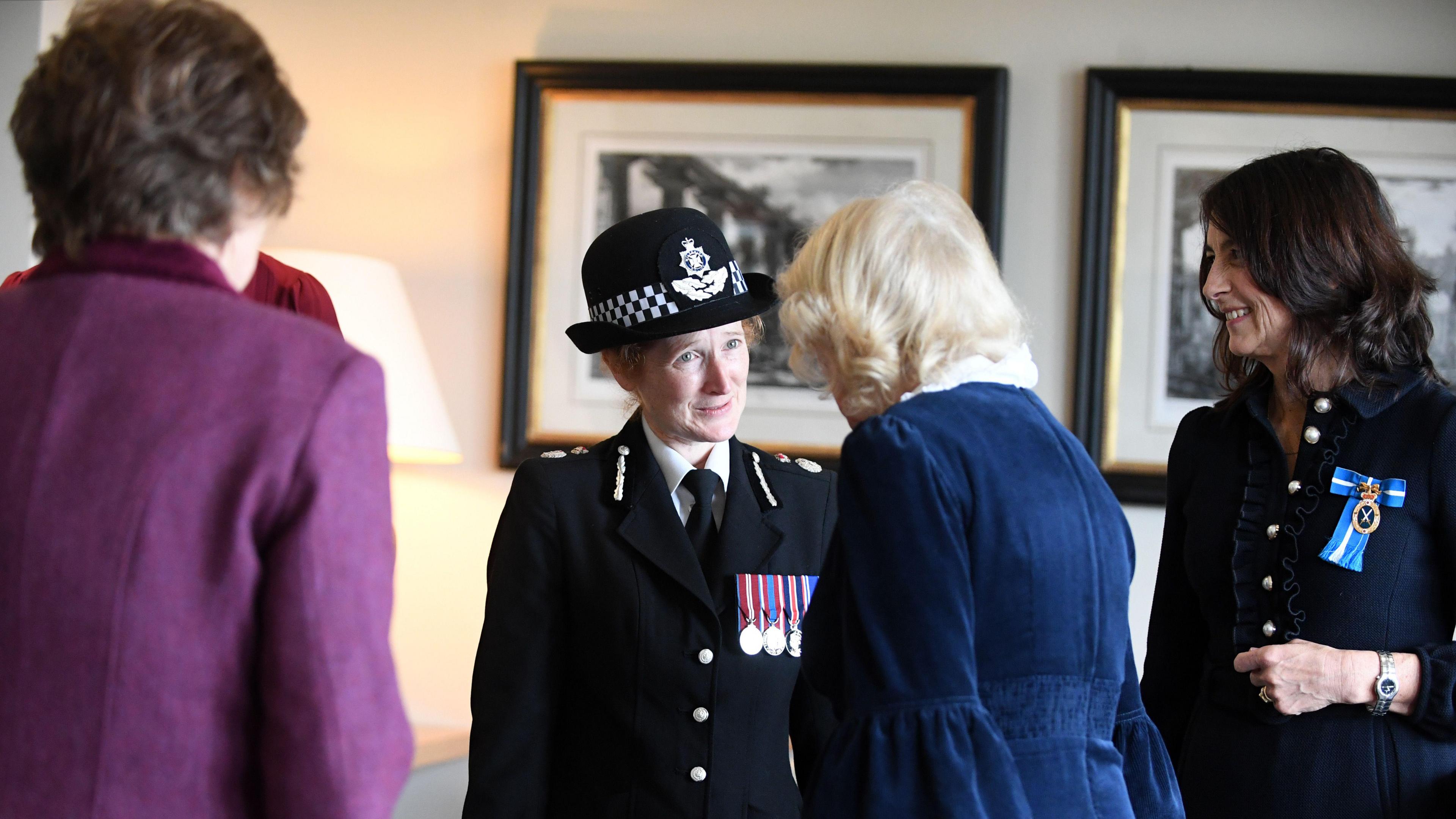 A ginger-haired woman in a police uniform chats with a woman with blonde hair (the Queen) with others looking on and picture frames in the background.