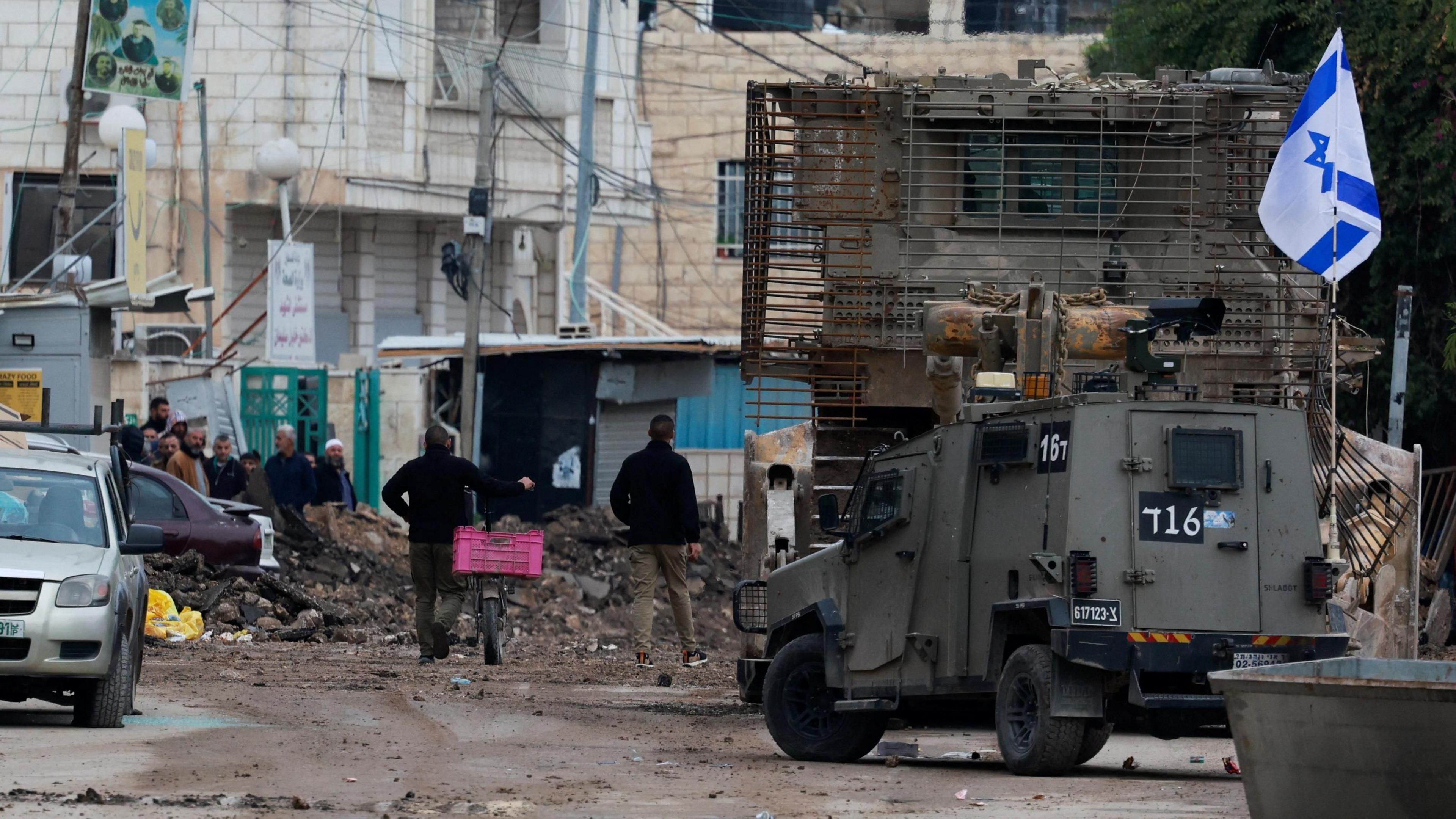 Palestinians walk past an Israeli military vehicle and an armoured bulldozer during a major operation by Israeli security forces in Jenin, in the occupied West Bank (22 January 2024)