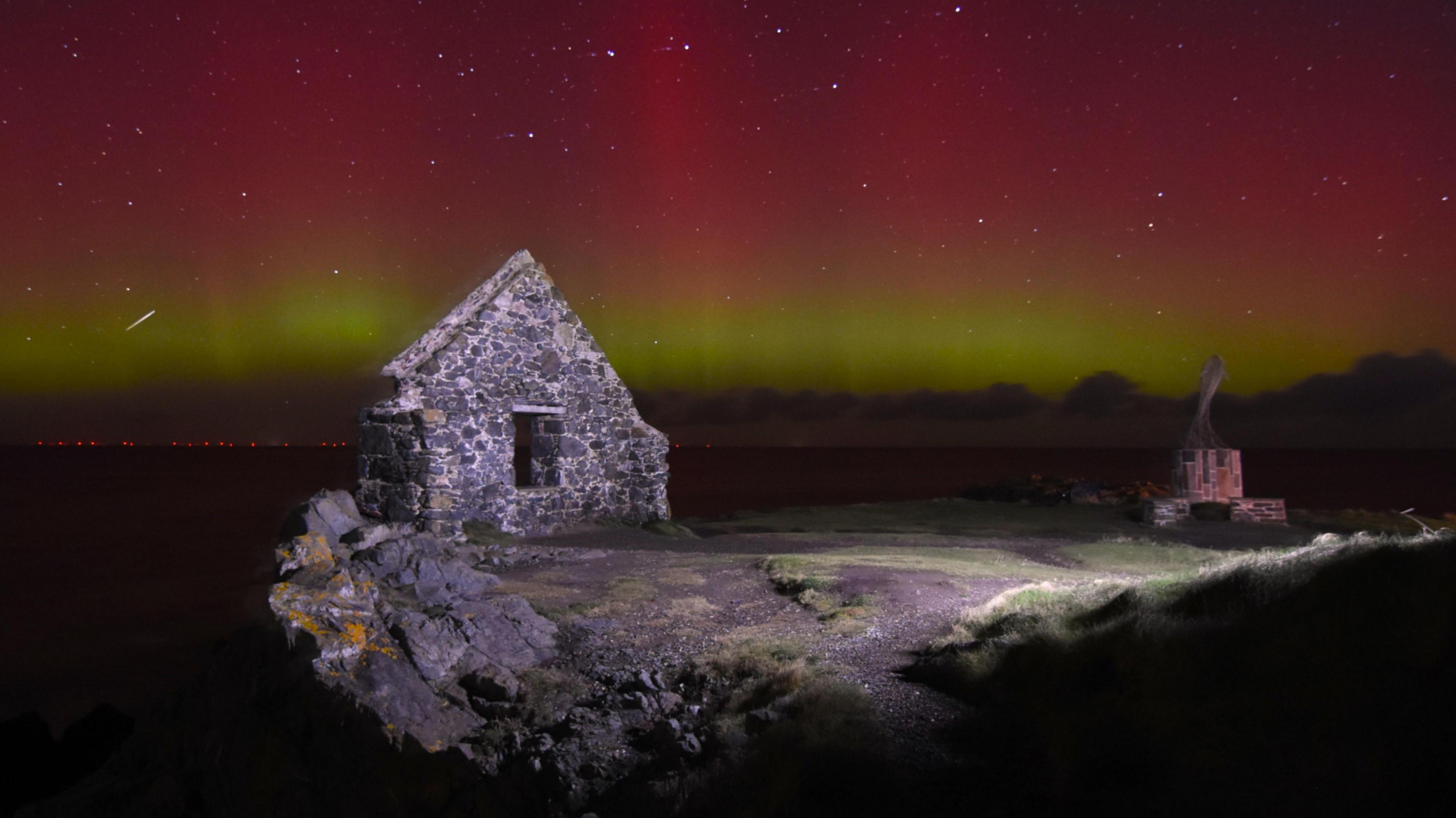 Portsoy Harbour at night with the Northern Lights overhead. The sky is multicoloured. There is a band of yellow-green light underneath a larger red glow. Several stars can be seen poking through the lights. One appears to be a shooting star which is brighter than the others. In the foreground, part of an abandoned building. It is shaped like the end of a house. A stone area can also be seen in the foreground. 