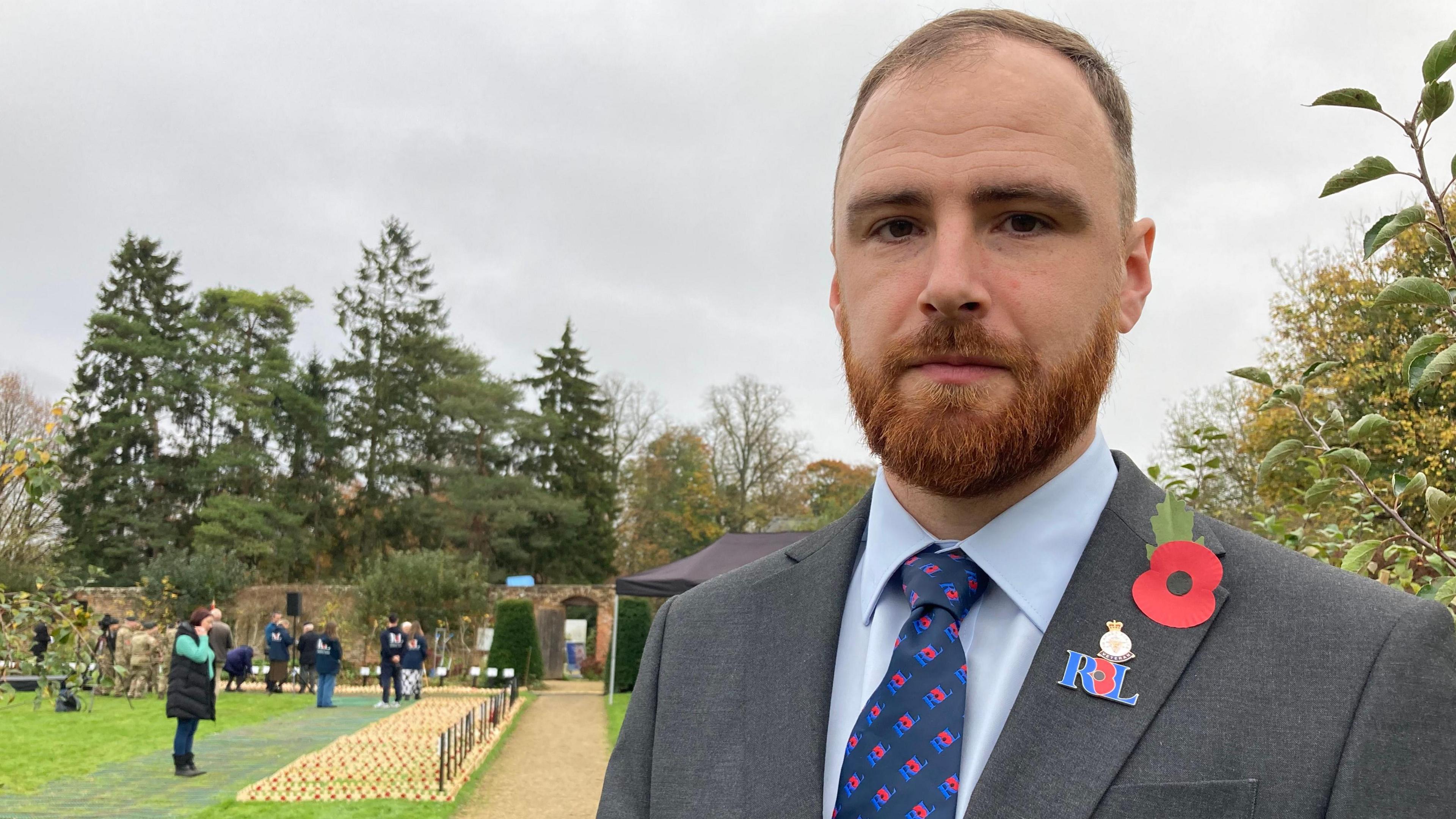 Peter Saunders of the Royal British Legion stands in the field of Remembrance on a cloudy day. People in the background can be seen looking at the memorials, in a walled garden surrounded by trees. Mr Saunders is wearing a dark grey blazer with a Royal British Legion badge and paper poppy on his lapel, and poppies on his tie. He has short brown hair and a ginger beard.