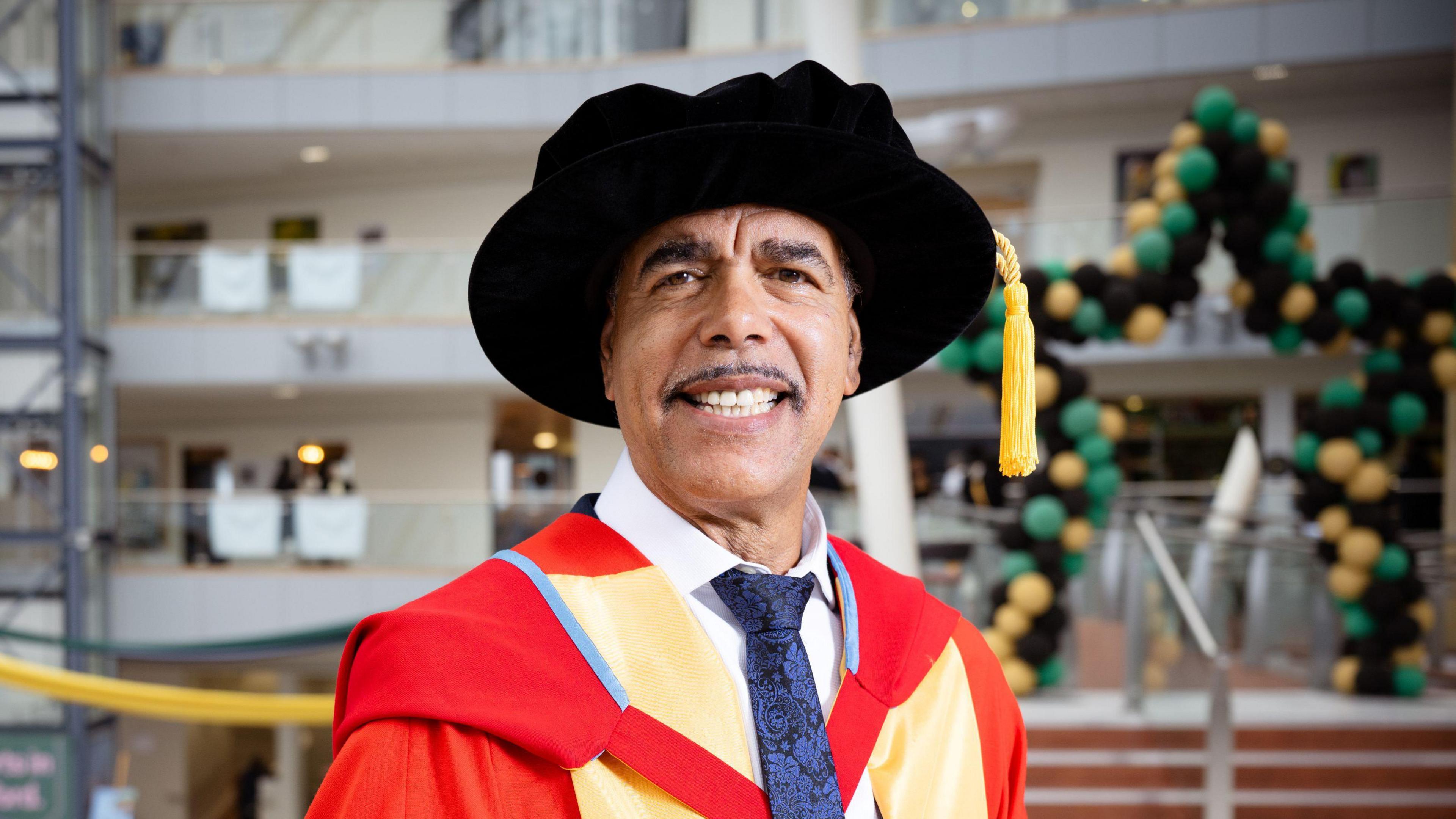 A man in a red and yellow robe, black hat and white shirt with blue tie. He is standing inside a university building smiling and looking at the camera