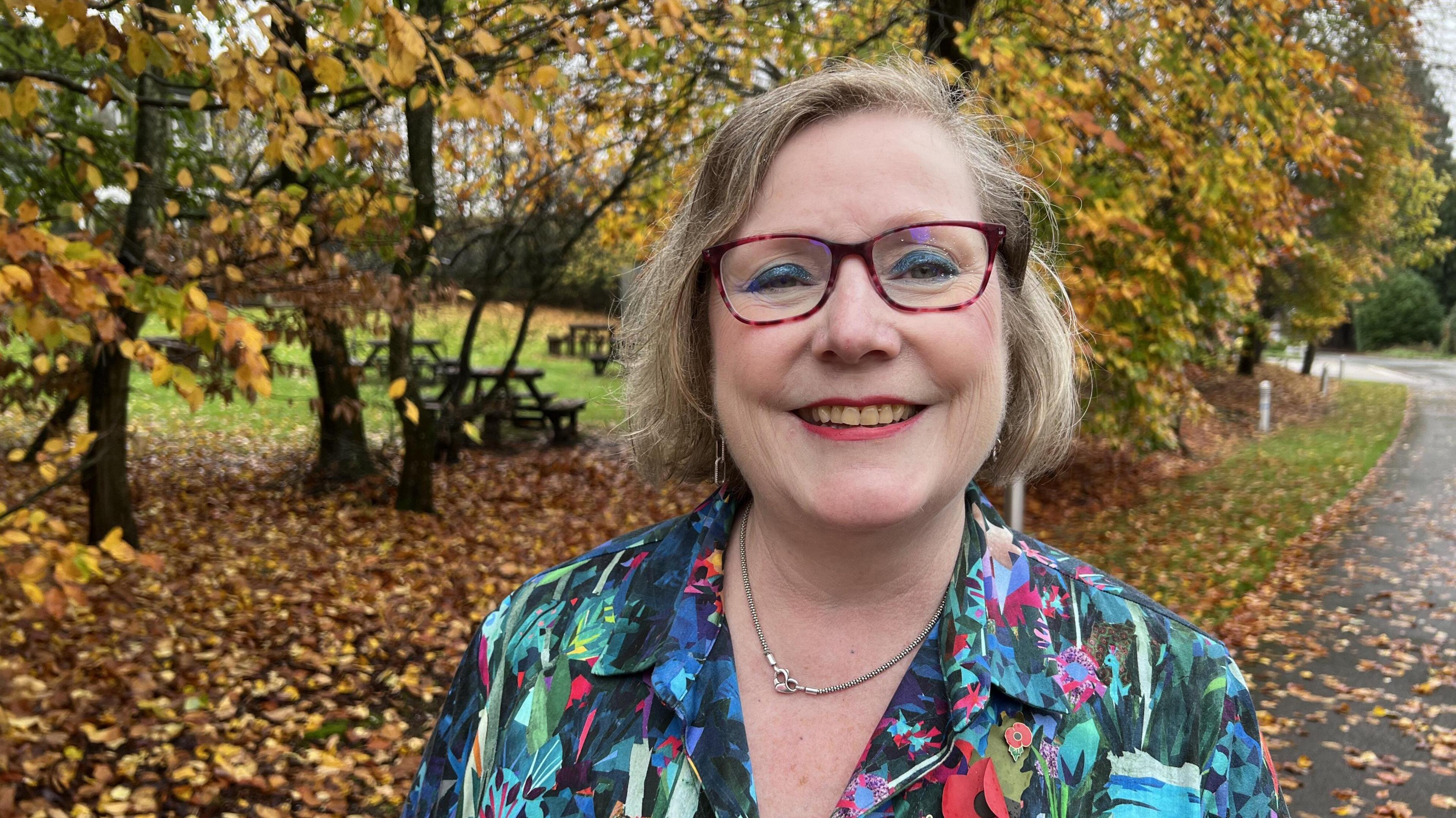 Woman smiling wearing glasses in front of autumnal trees. She wears a flowery jacket and glasses with bob length blond hair.