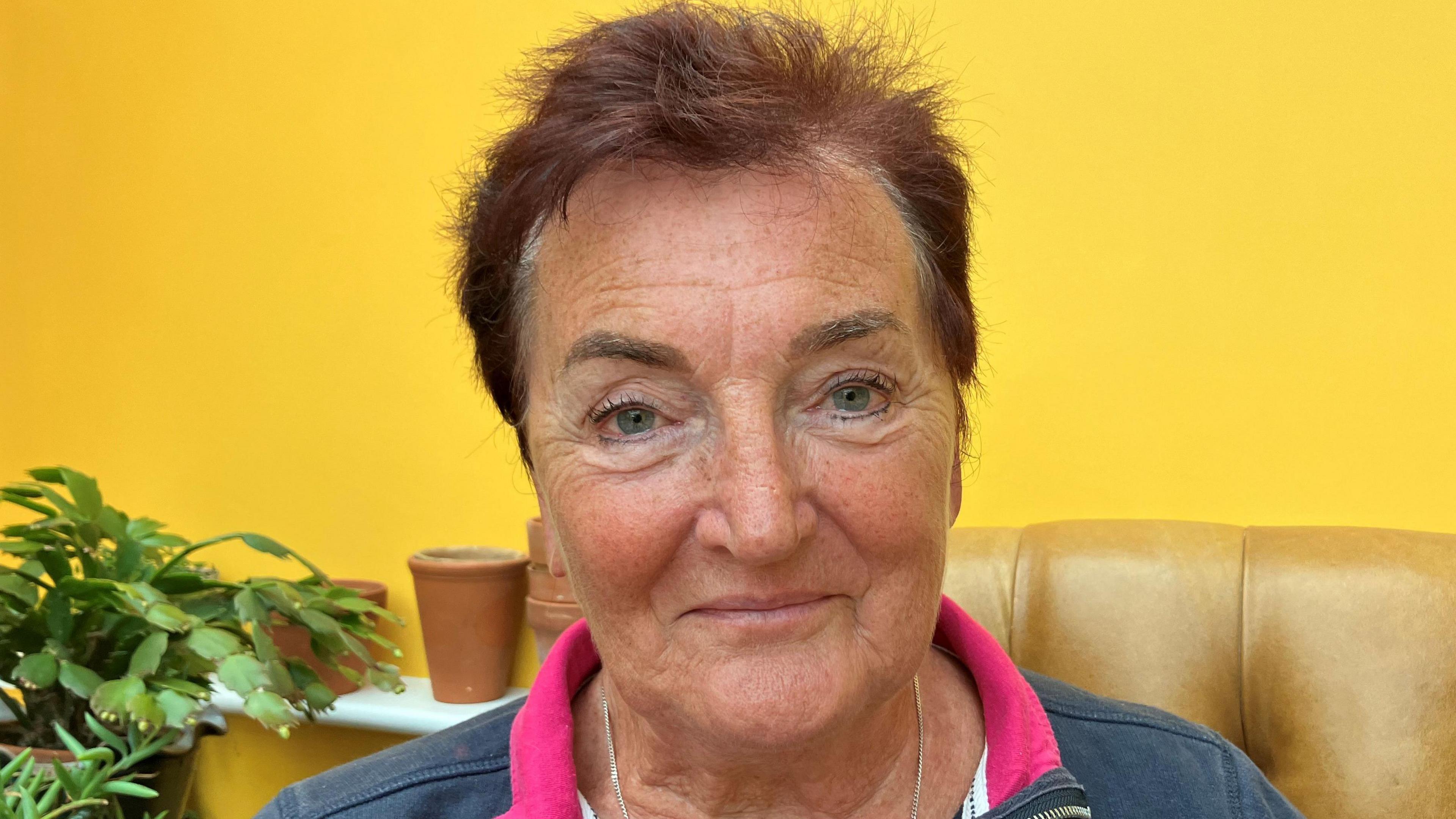 Sally Nicholls, a woman with short reddish hair, sits on a leather chair against a backdrop of yellow wallpaper