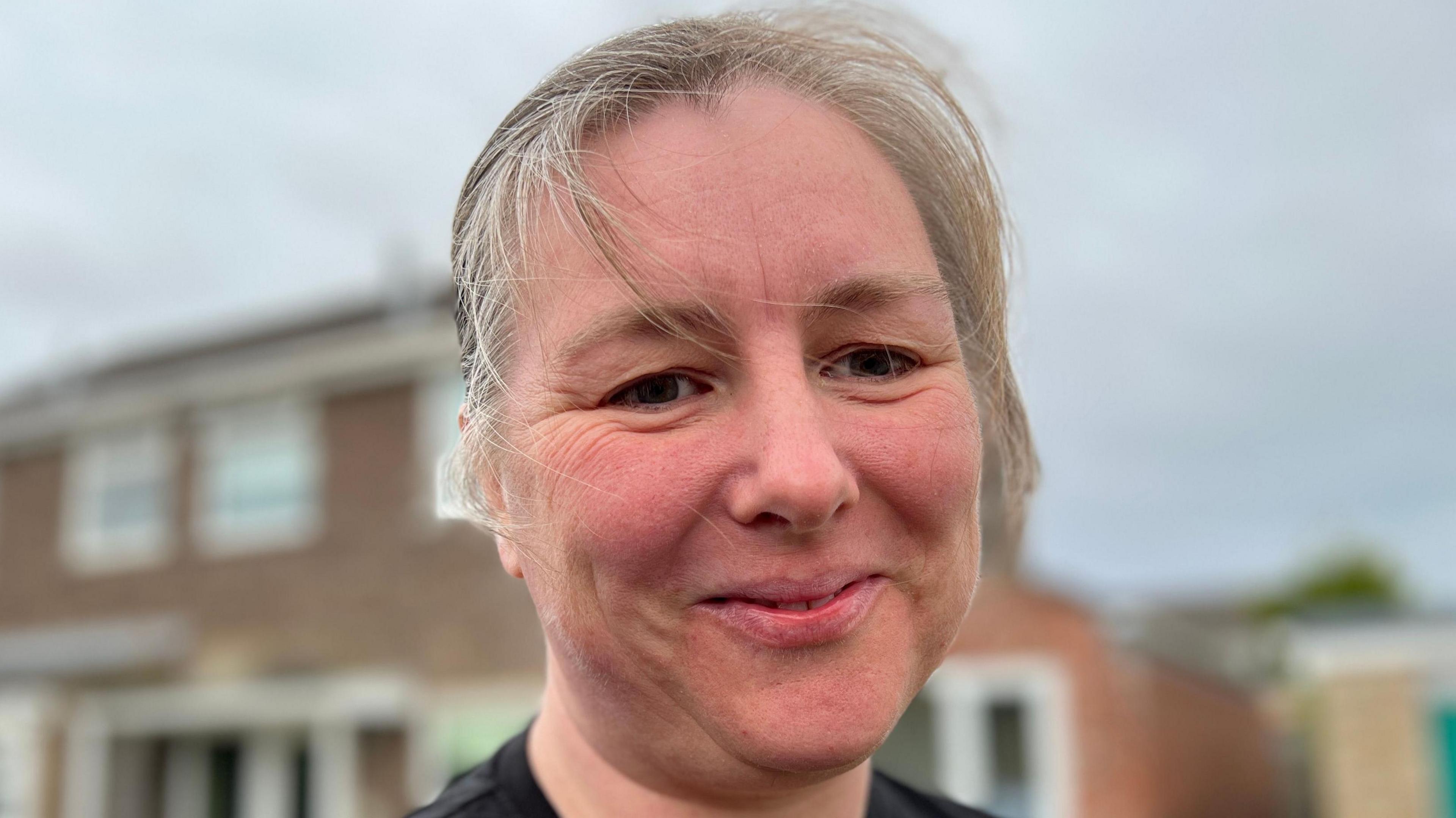 Sally Griffen smiling at the camera, she has grey hair and a house is in the distance behind her