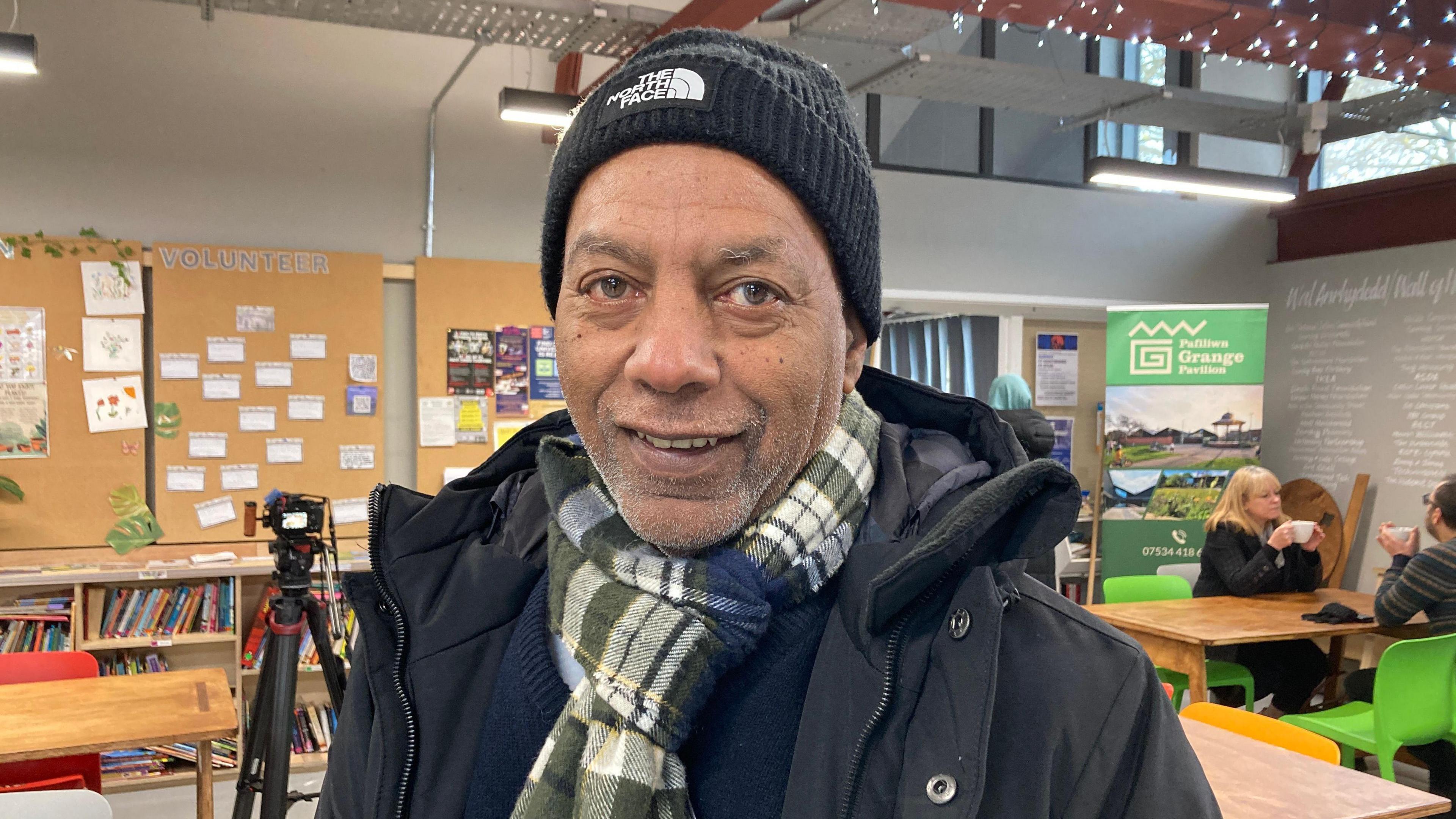 Steve Khaireh in a North Face woolly hat and green, white and blue check scarf at a community centre in Grangetown. A camera on a tripod is behind him and two people are having a hot drink at a table over his left shoulder 