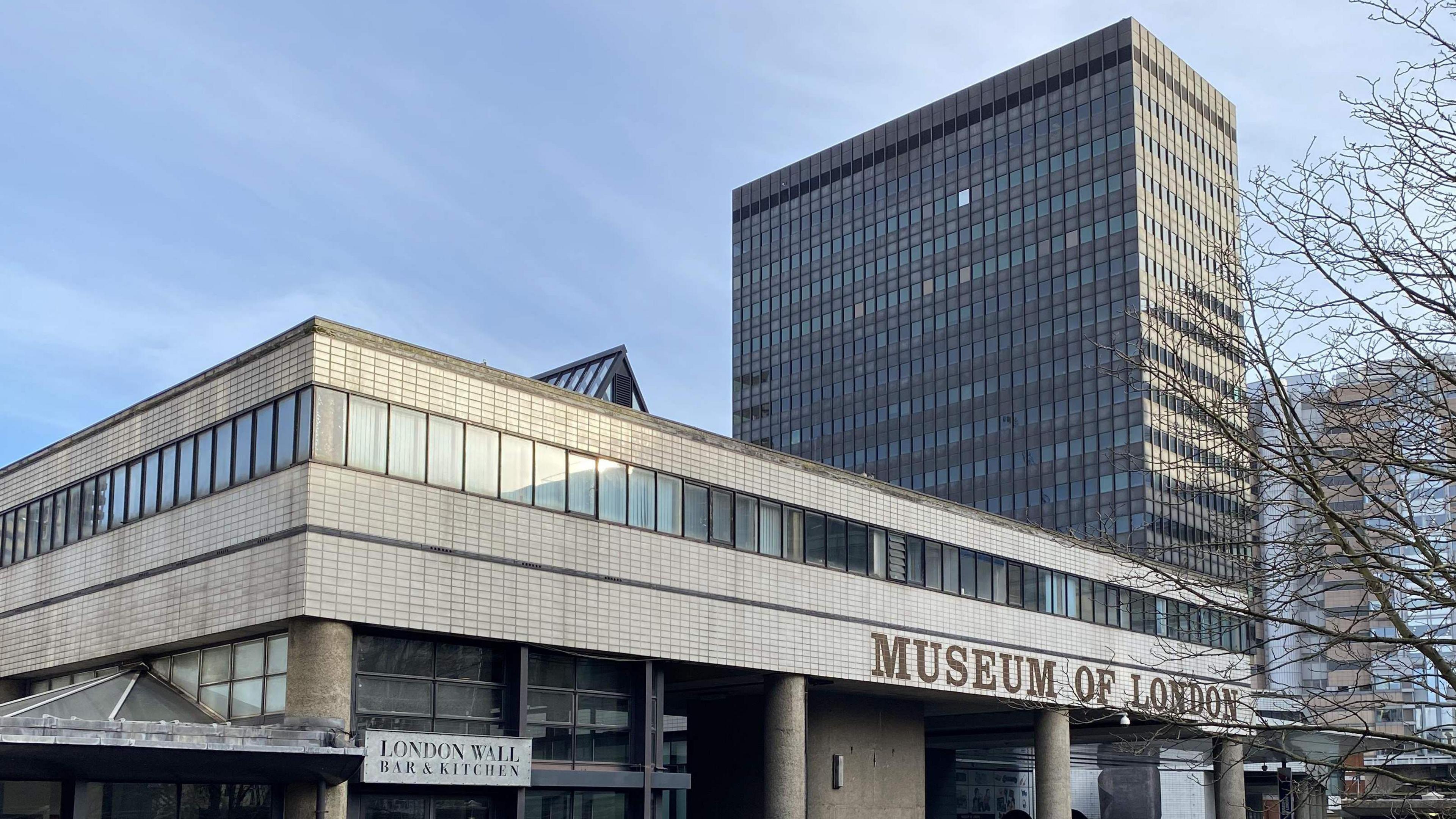 Front view of the former building of the Museum of London and the office block Bastion House right behind it. The former museum building is a wide concrete block sitting on top of concrete pillars - four of which are in view, and has 'Museum of London' written across the front. Bastion House is a 17-storey office block in the shape of a standard skyscraper, grey in colour.