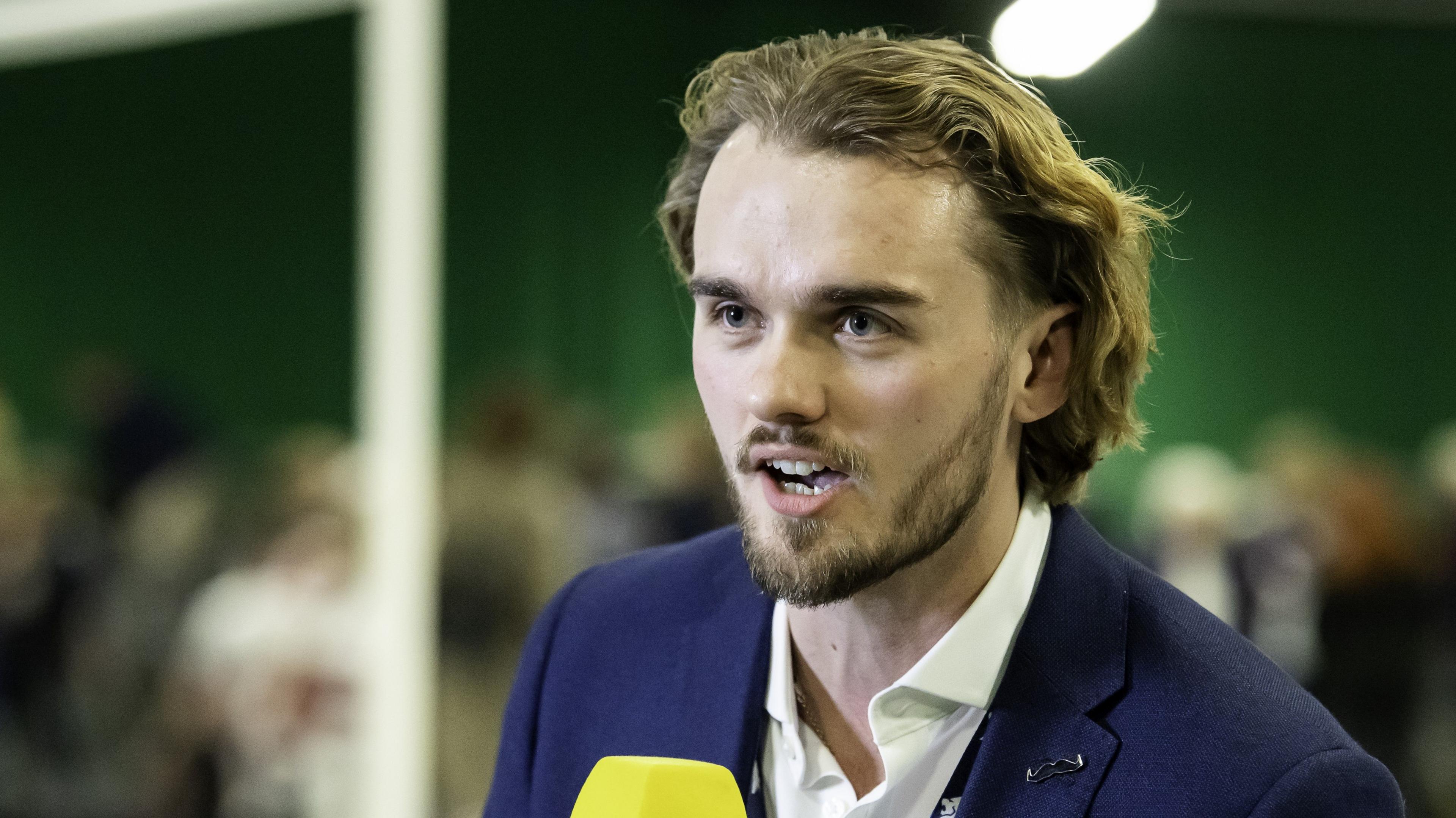 Tom Coyd looks into the distance. He is holding a microphone and speaking