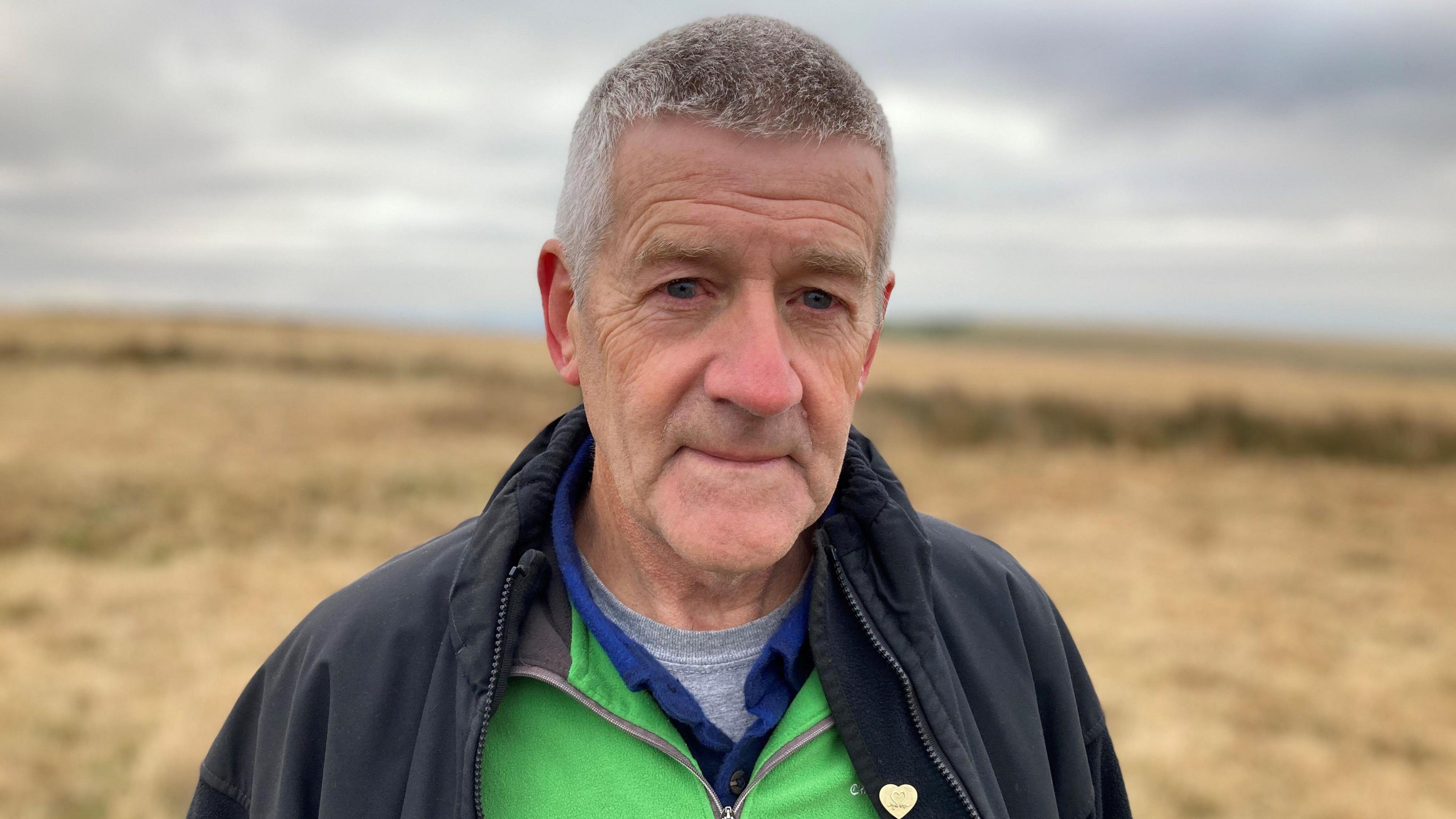 Gareth Richards of Mawr Community Council stands in front of the Tor Clawdd ring cairn wearing a black coat.  The banks of the prehistoric monument are visible behind him.  