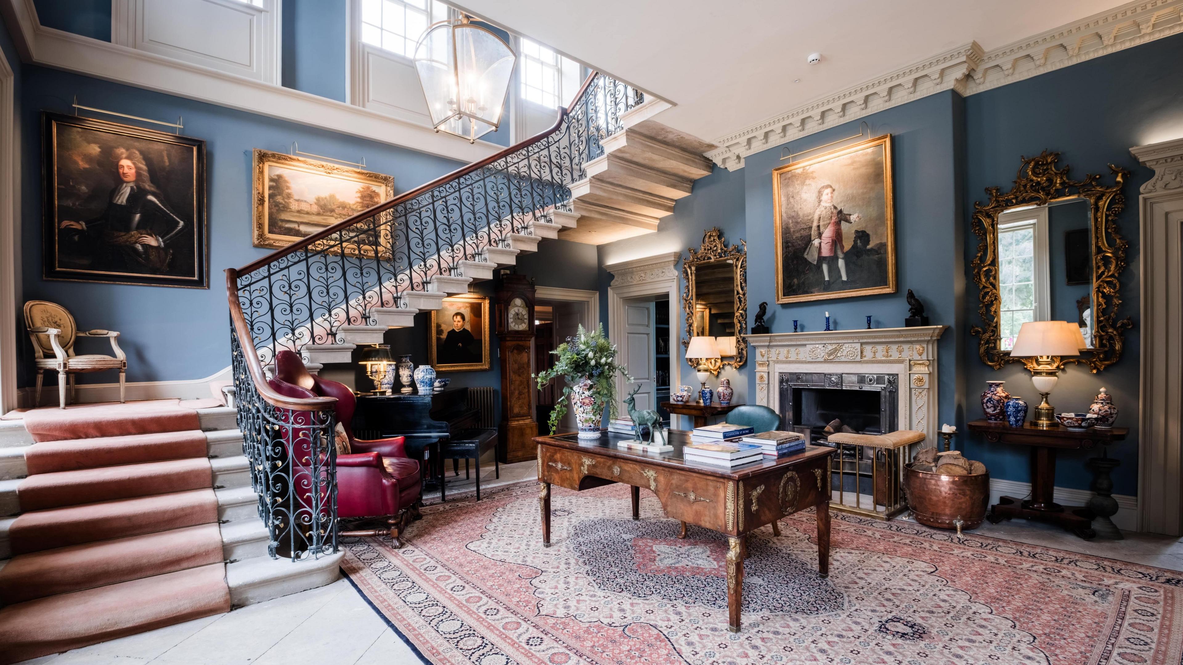 The grand entrance to Hotham Hall. A tradition fireplace is surrounded by oil paintings and antique furniture. The walls are blue with a stone floor covered with a blue, pink and cream rug.