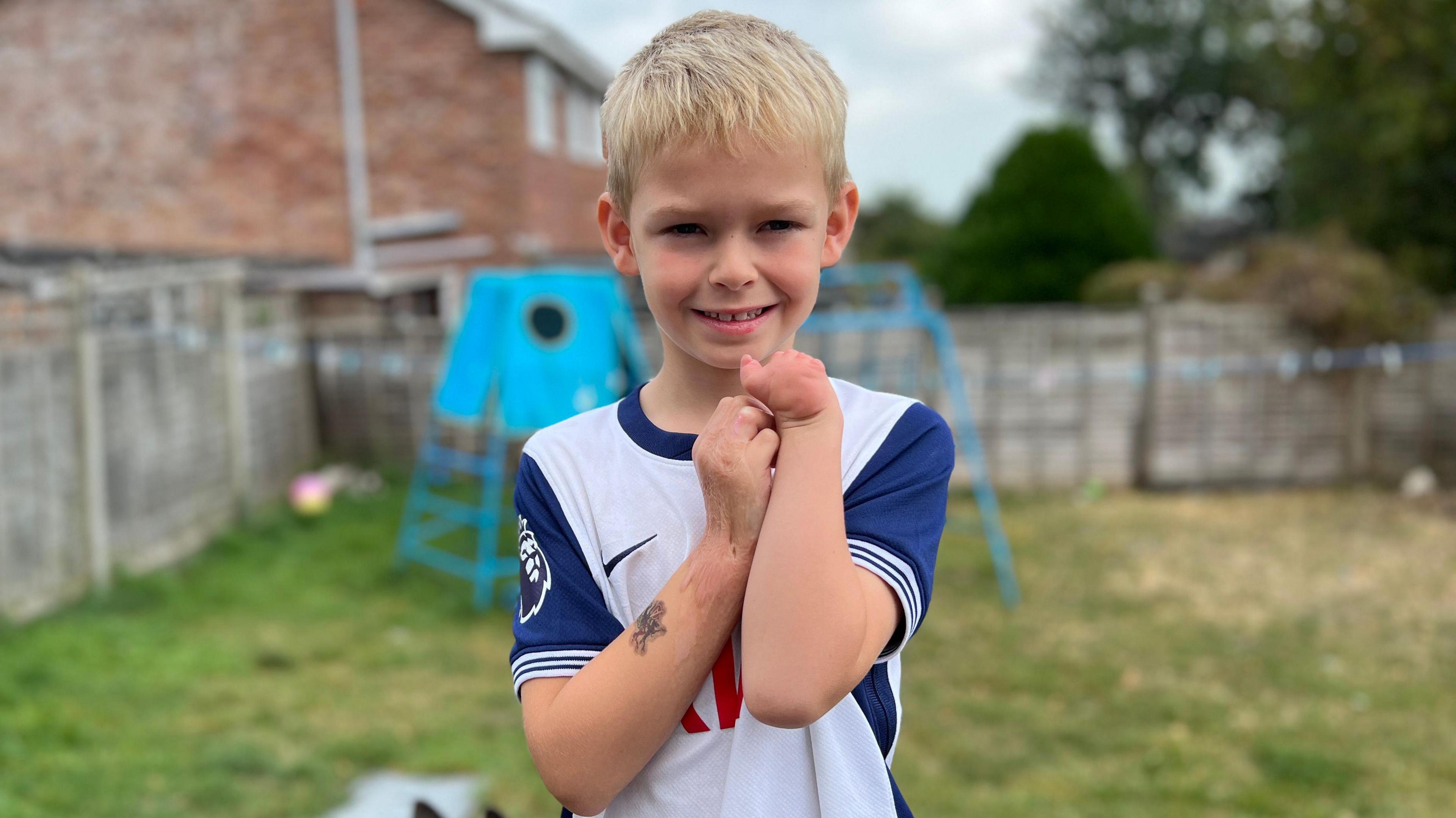 Brodie in his back garden. He is blonde and wearing a white top with blue sleeves. He is holding his arms over his chest. He only has one hand - which is something he was born with. He is looking at the camera and smiling.