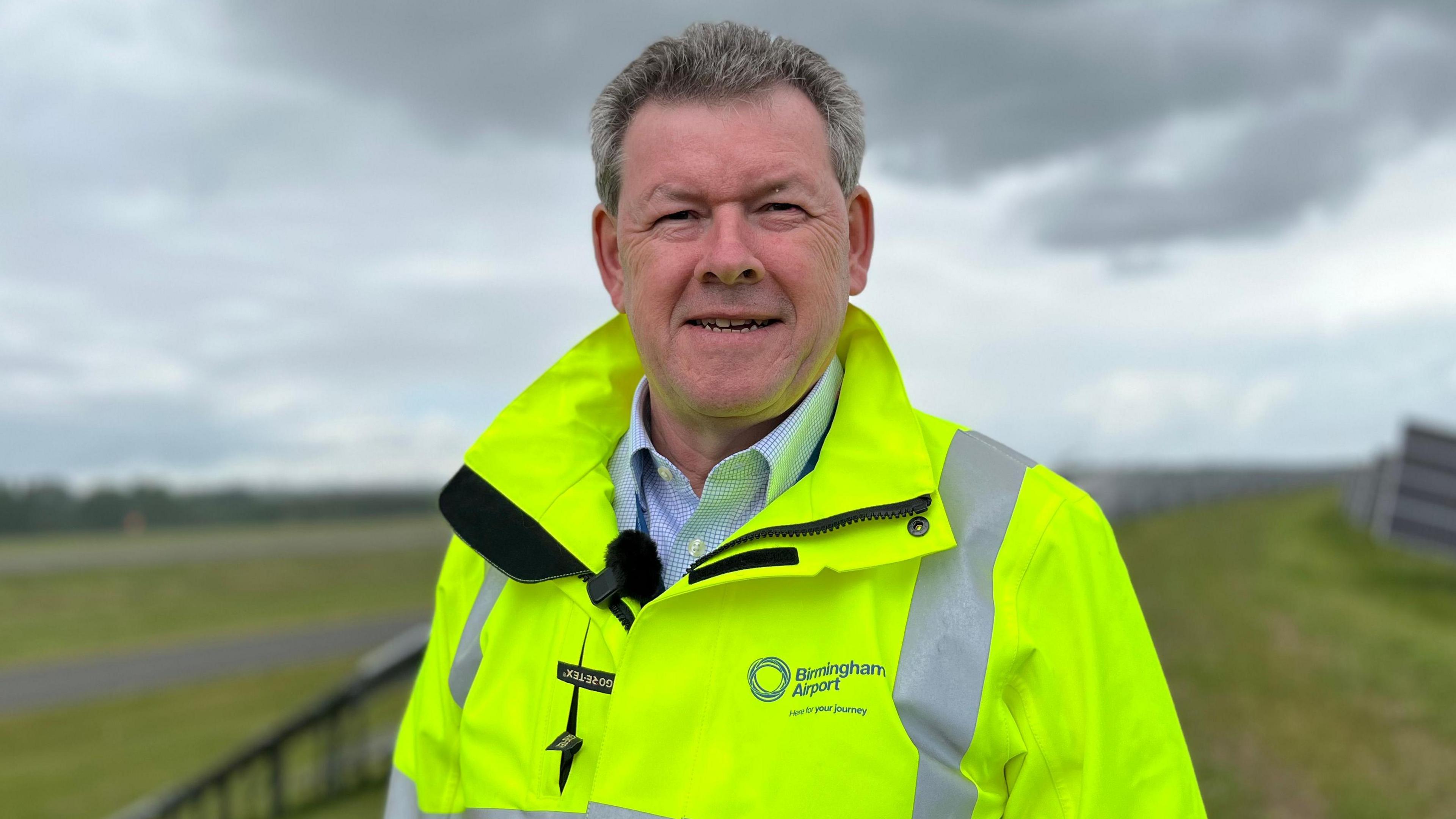 A man is wearing a yellow high-vis jacket and is standing in a field next to an airport runway. There are solar panels installed on the field.