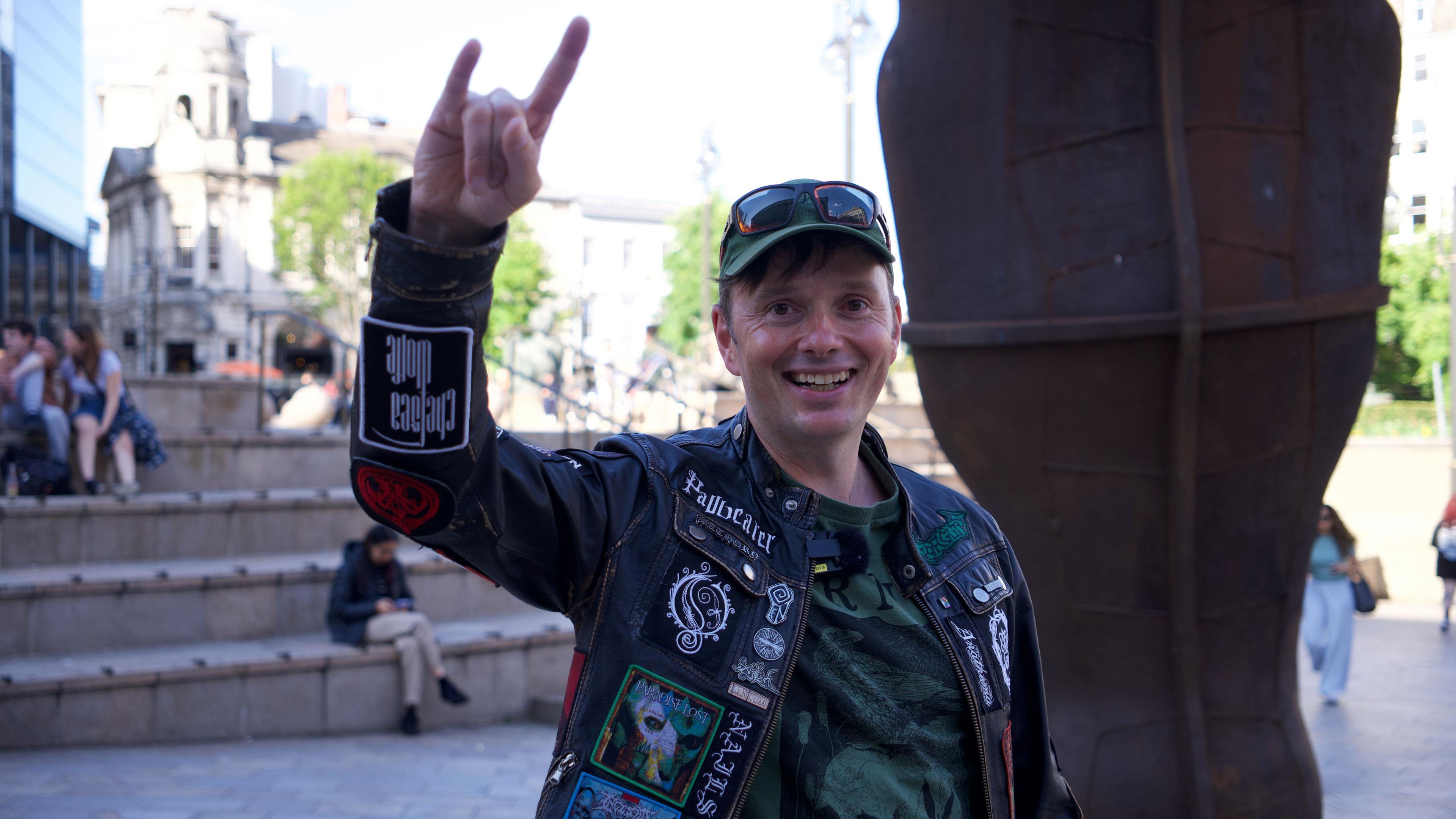 A man in a black jacket covered in band logos wears a green baseball hat with sunglasses balanced top. He is making a heavy metal sign with his index and little fingers next to Birmingham's Iron: Man statue.