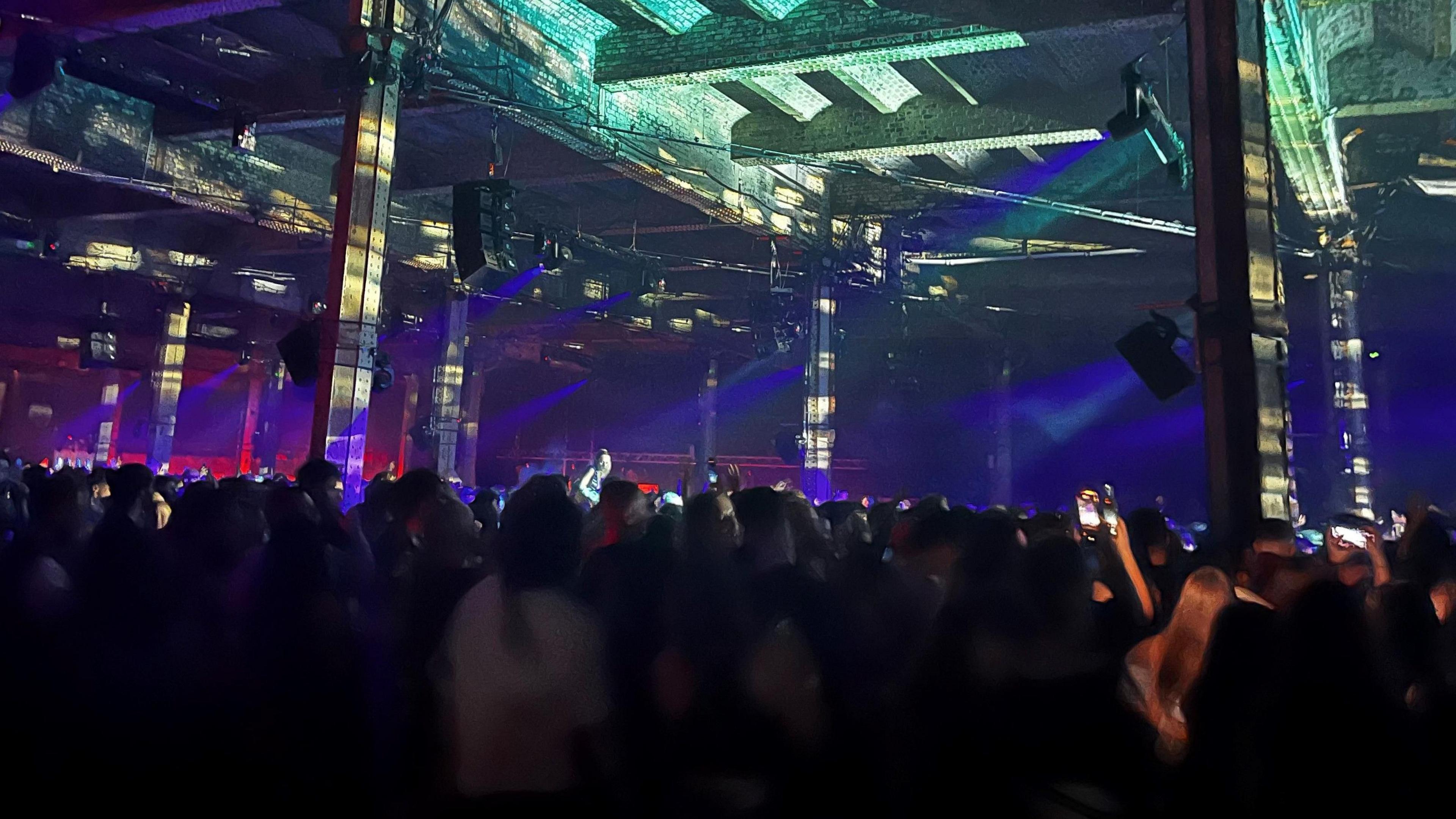 A nightclub with a crowd looking ahead to the DJ booth. Strobe lighting beams across the crowds