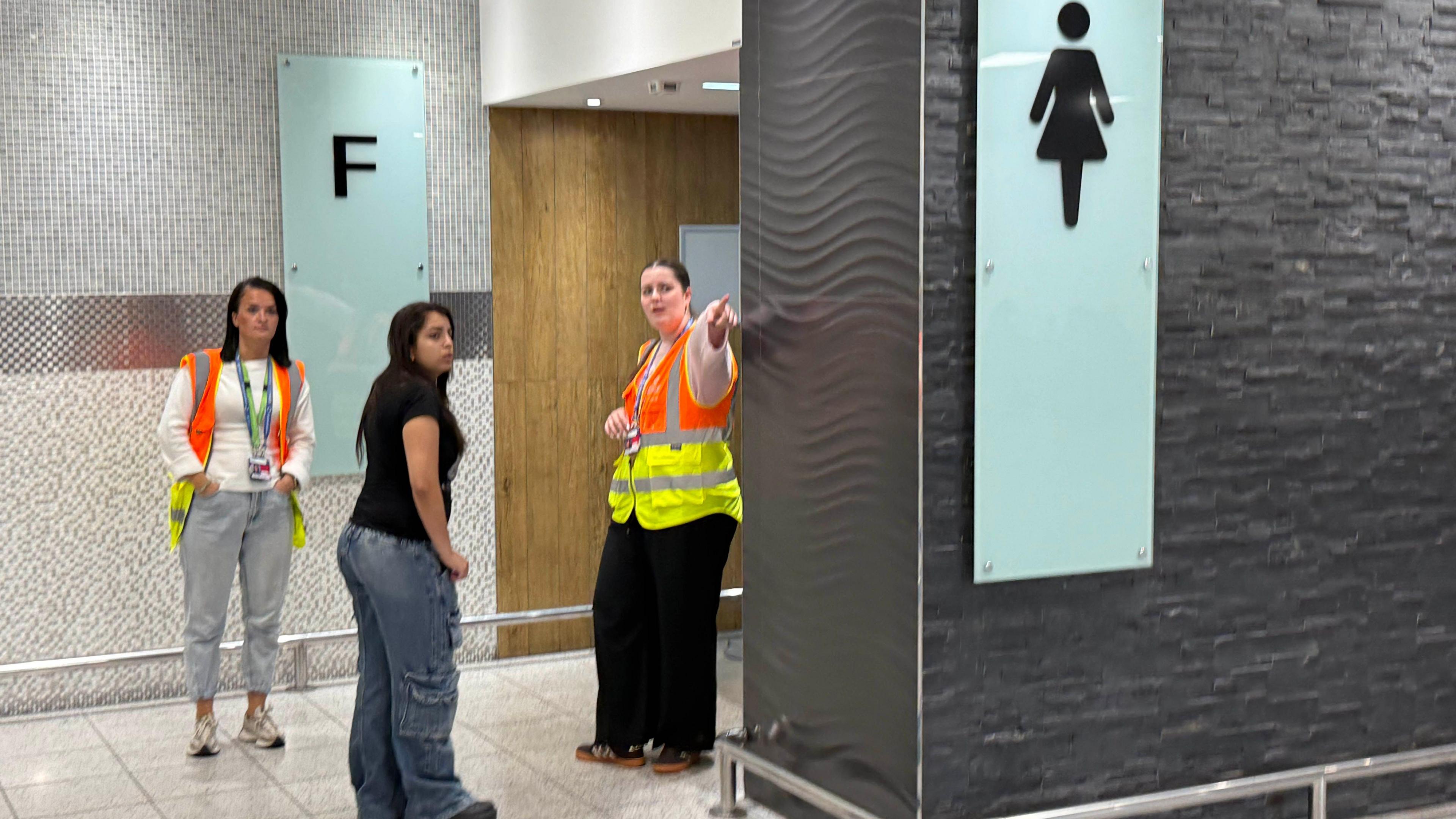 Two women in high vis coats at the entrance to a women's toilet. One of them is pointing another woman in the opposite direction.
