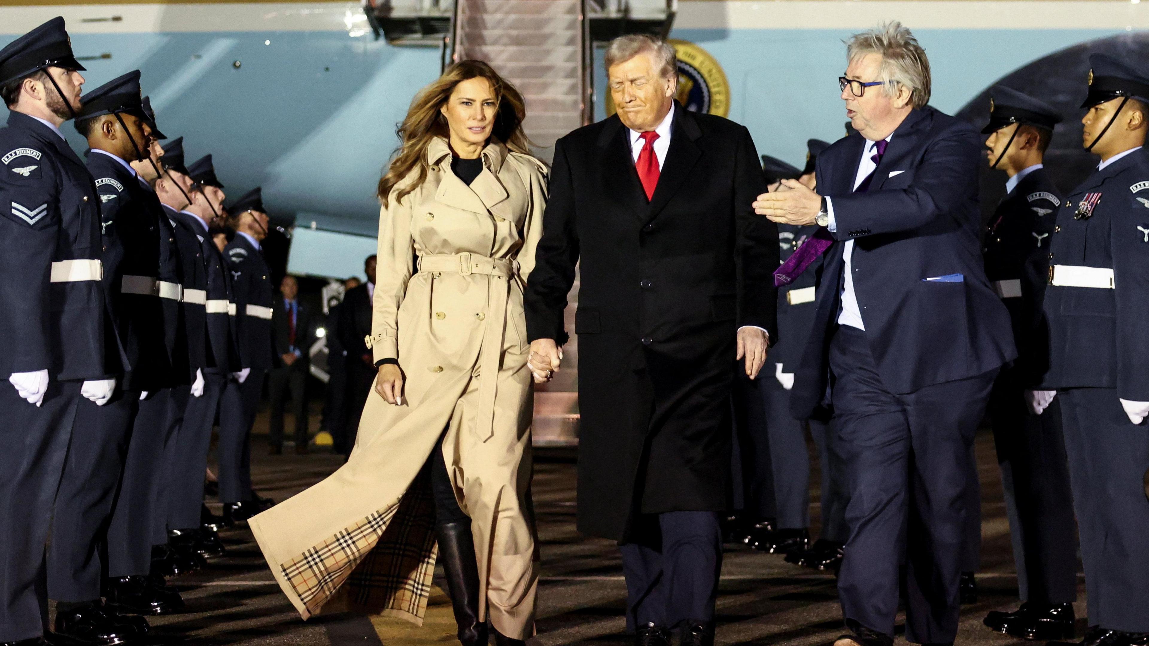 Donald and Melania Trump walk from Air Force One through a line of men in military uniform. Viscount Henry Hood walks alongside the pair.