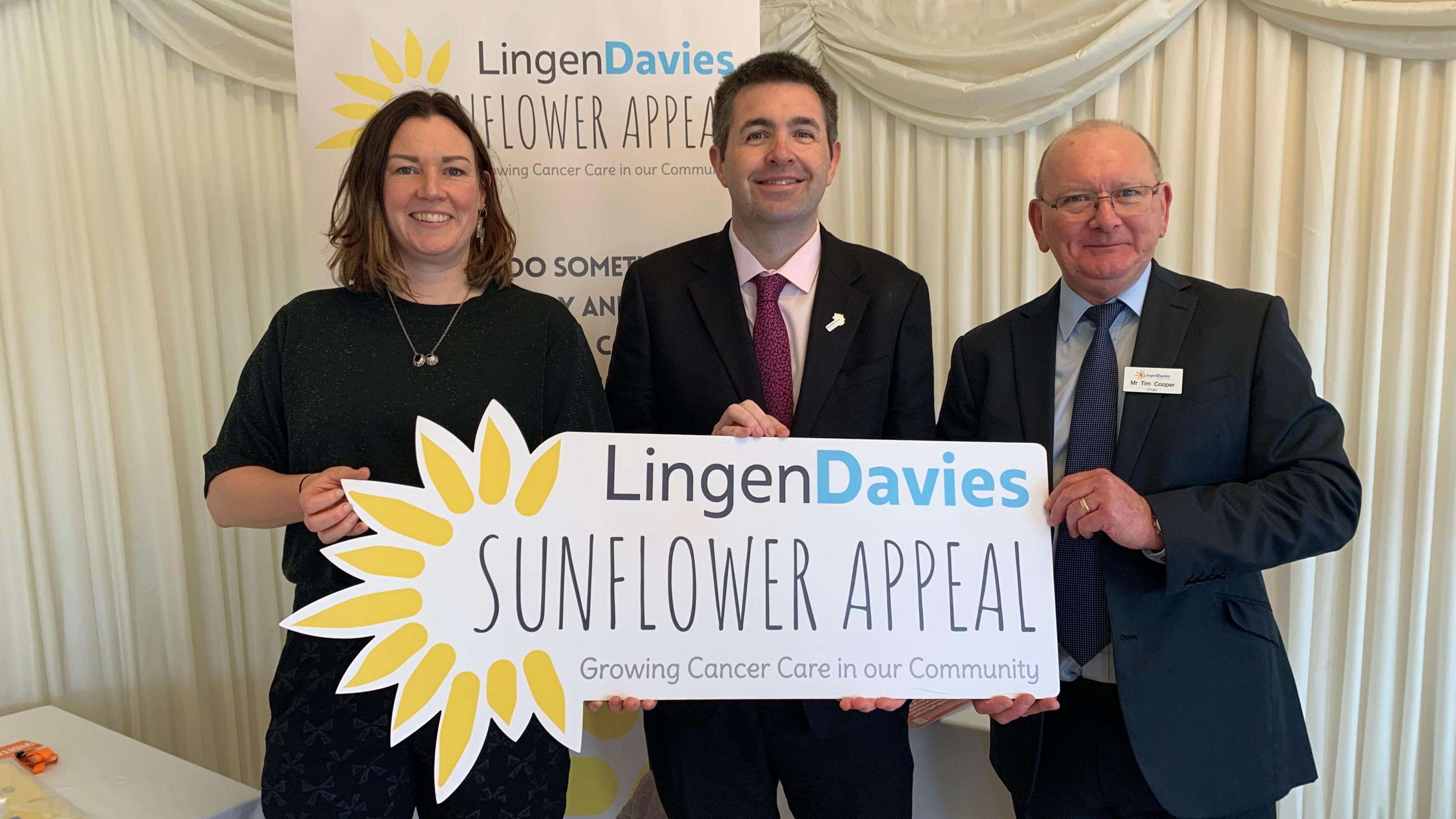 A woman and two men sanding in front of a white curtain and a sign reading "Lingen Davies Sunflower Appeal", and holding another sign saying the same thing with a yellow sunflower on it.