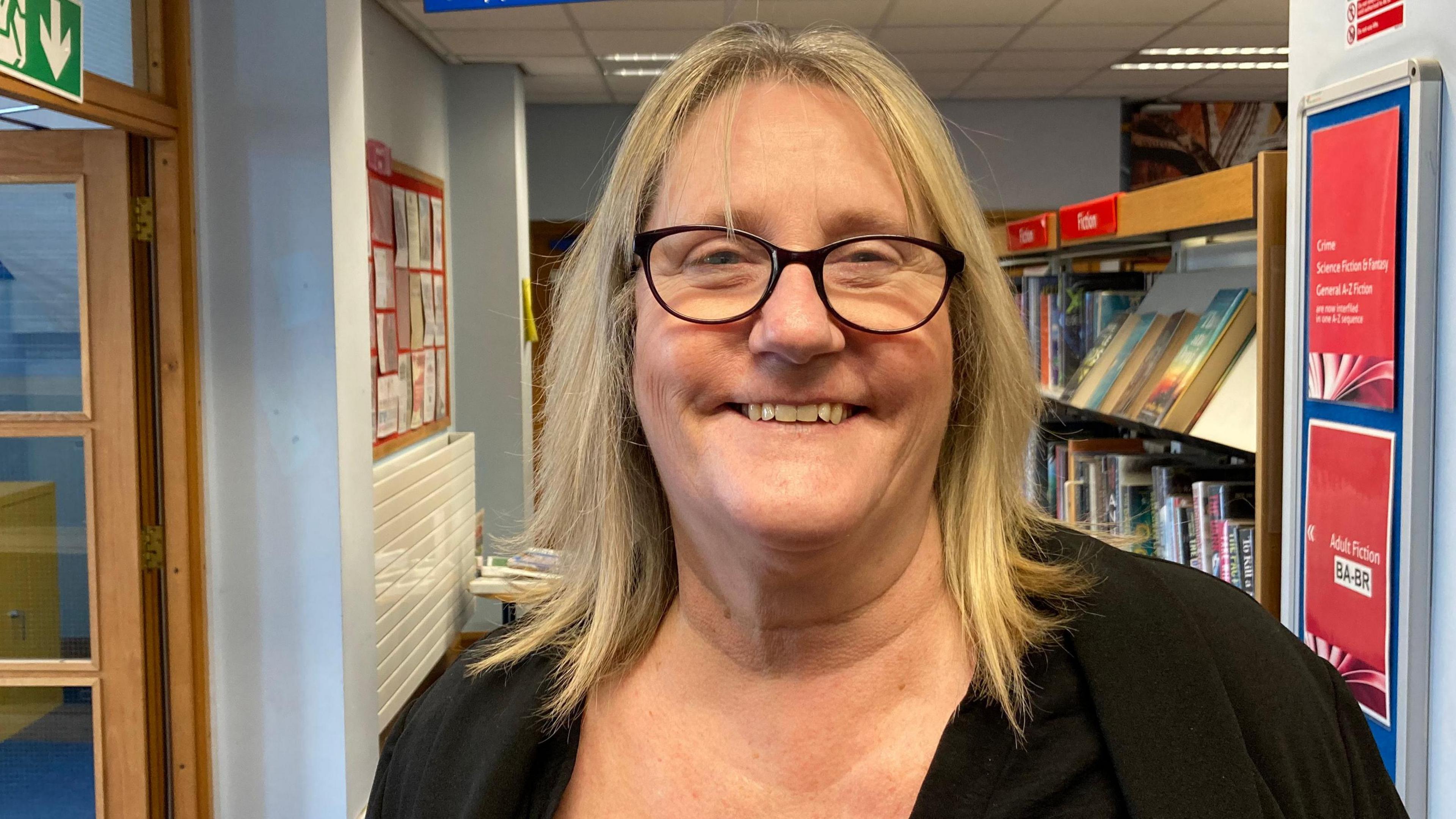 A woman wearing a black jumper and black glasses with blonde hair standing in front of some books