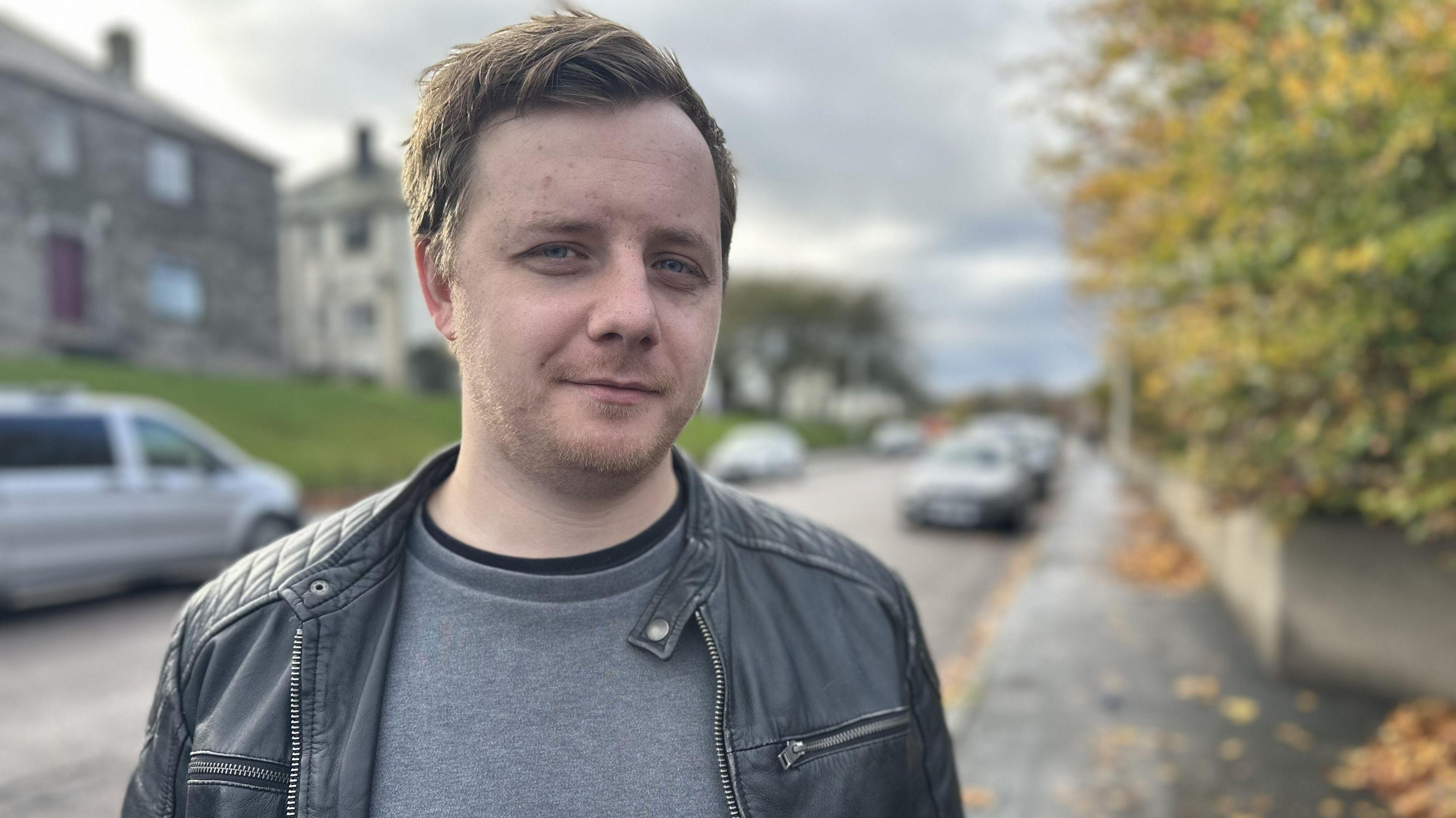 A man with dark hair wearing a grey sweater and black leather jacket. He is standing outdoors on a wet pavement beside a leafy green hedge. There are homes and parked cars in the background