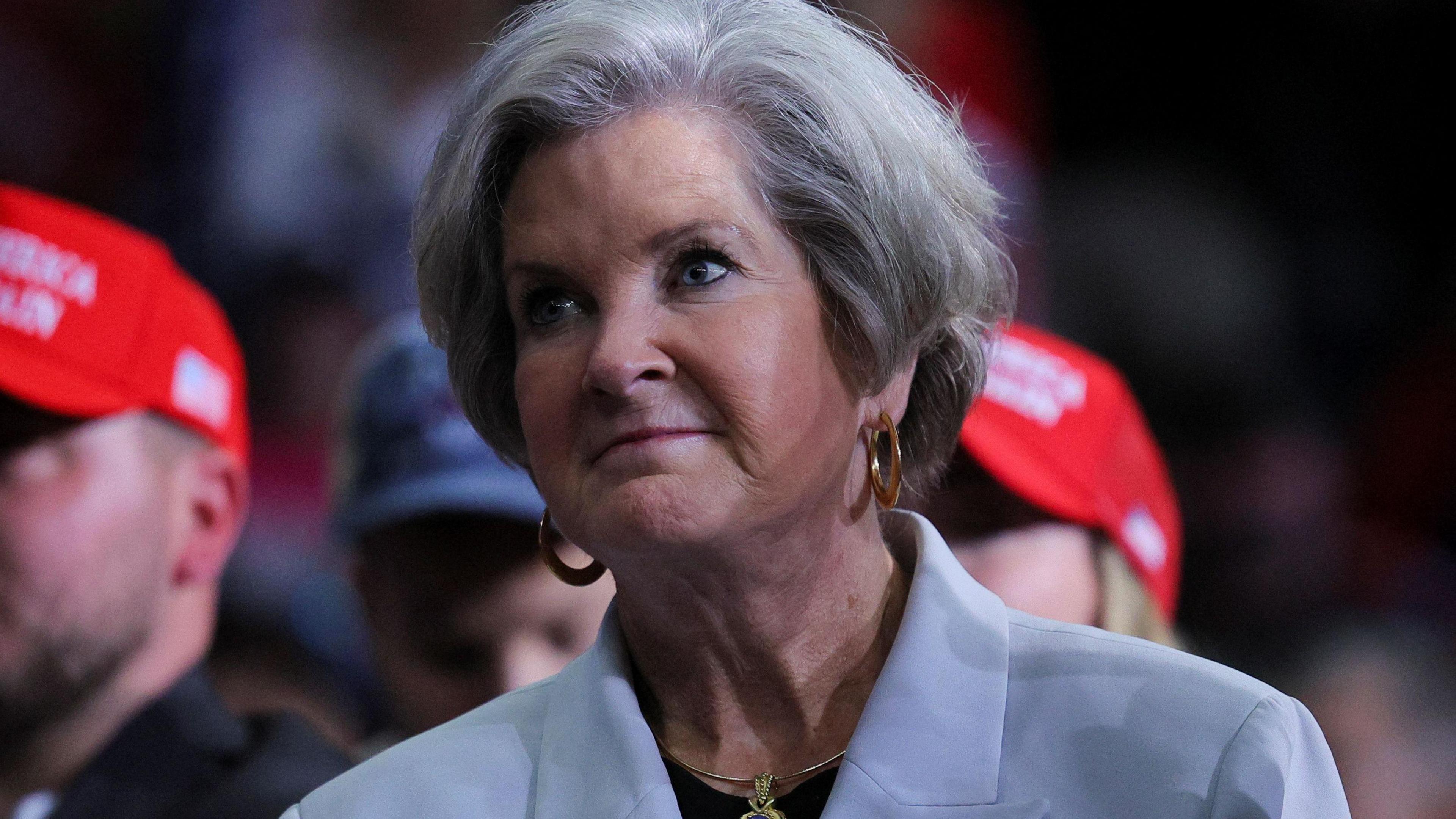 Susie Wiles, who has a grey cropped haircut, wears gold hoop earrings with a gold pendant necklace and a black top underneath a baby blue blazer which has a white and gold brooch on it as she looks on during a Trump rally